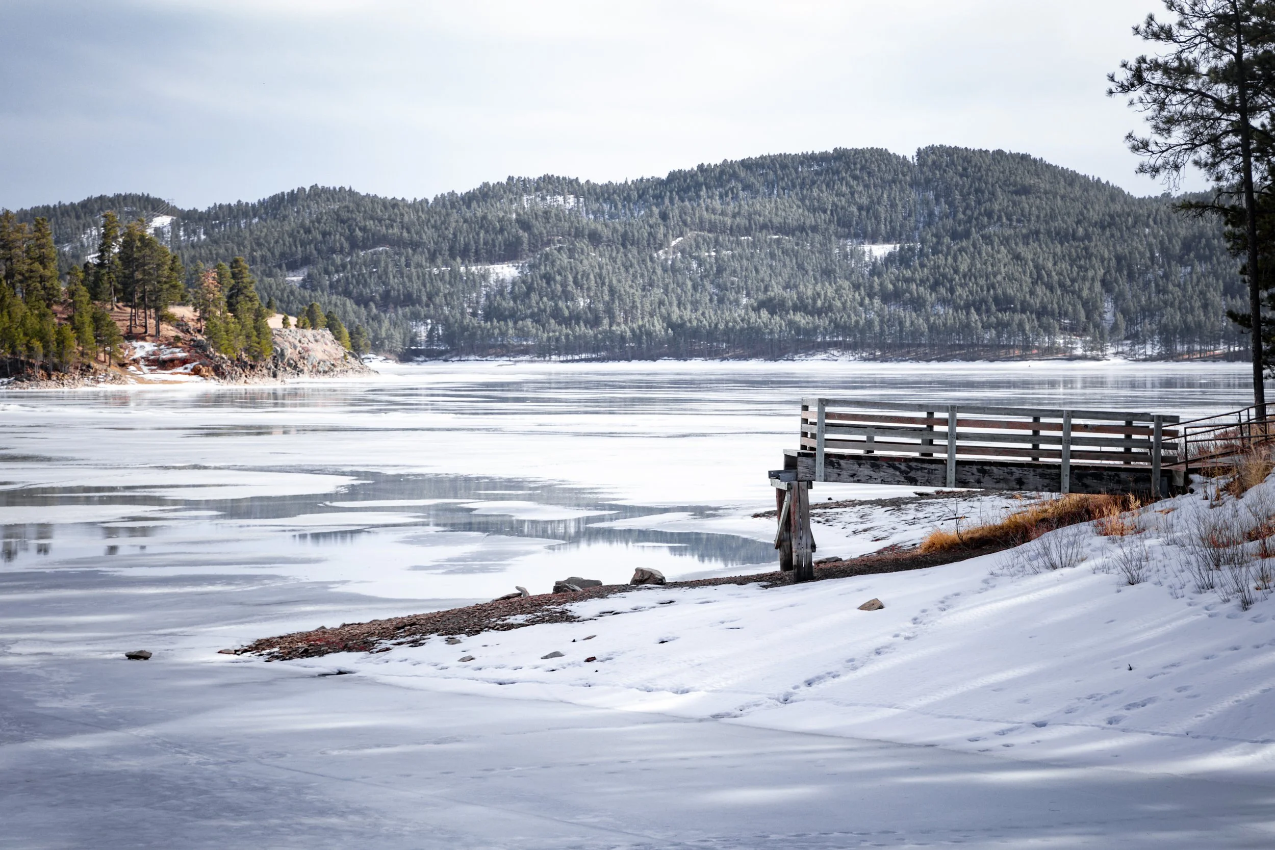 Snow-covered shoreline with a partially frozen lake and a wooden dock, surrounded by pine trees and mountains in the background.