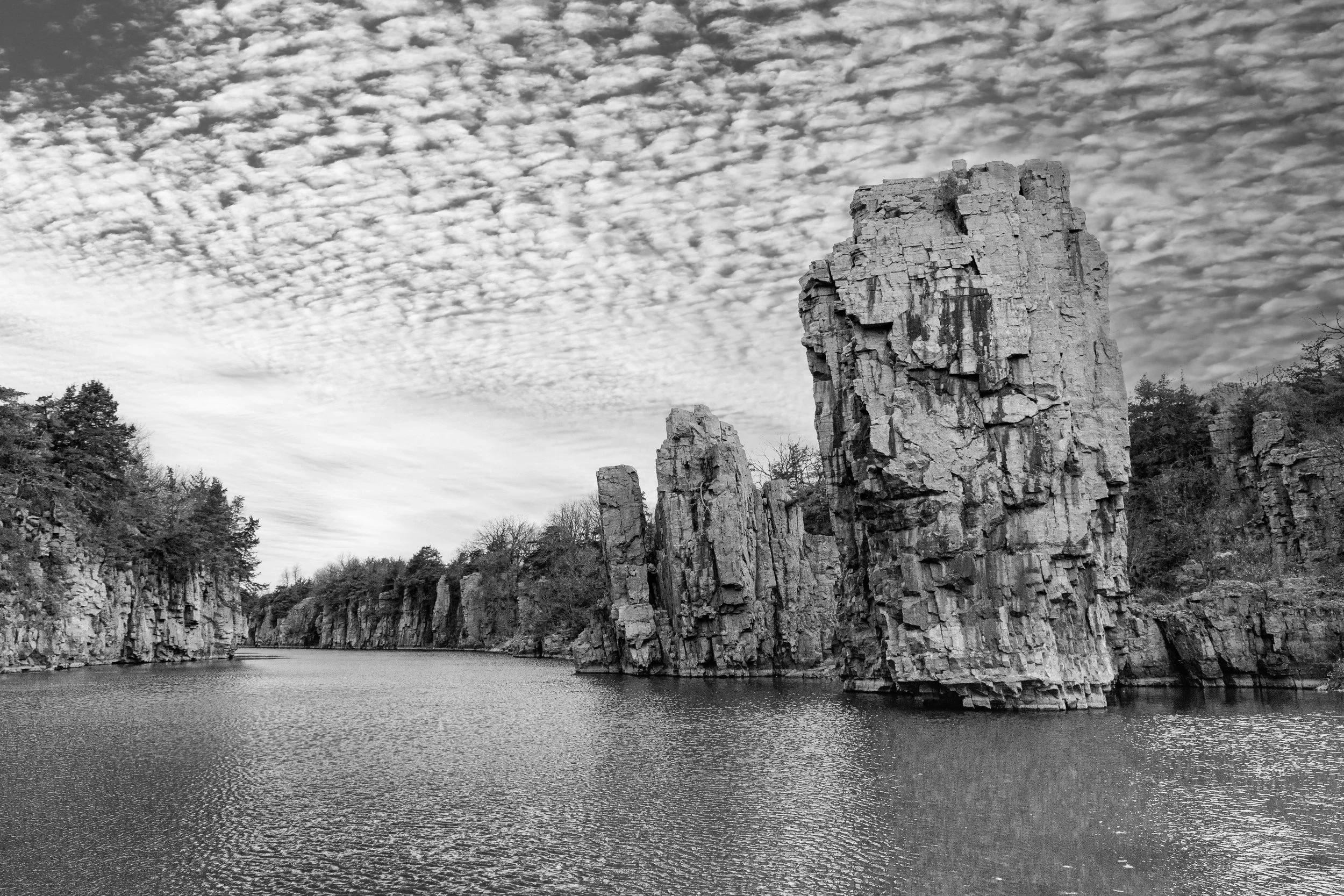 Black and white photo of a body of water with tall, rugged rock formations along the shoreline and a cloudy sky above.
