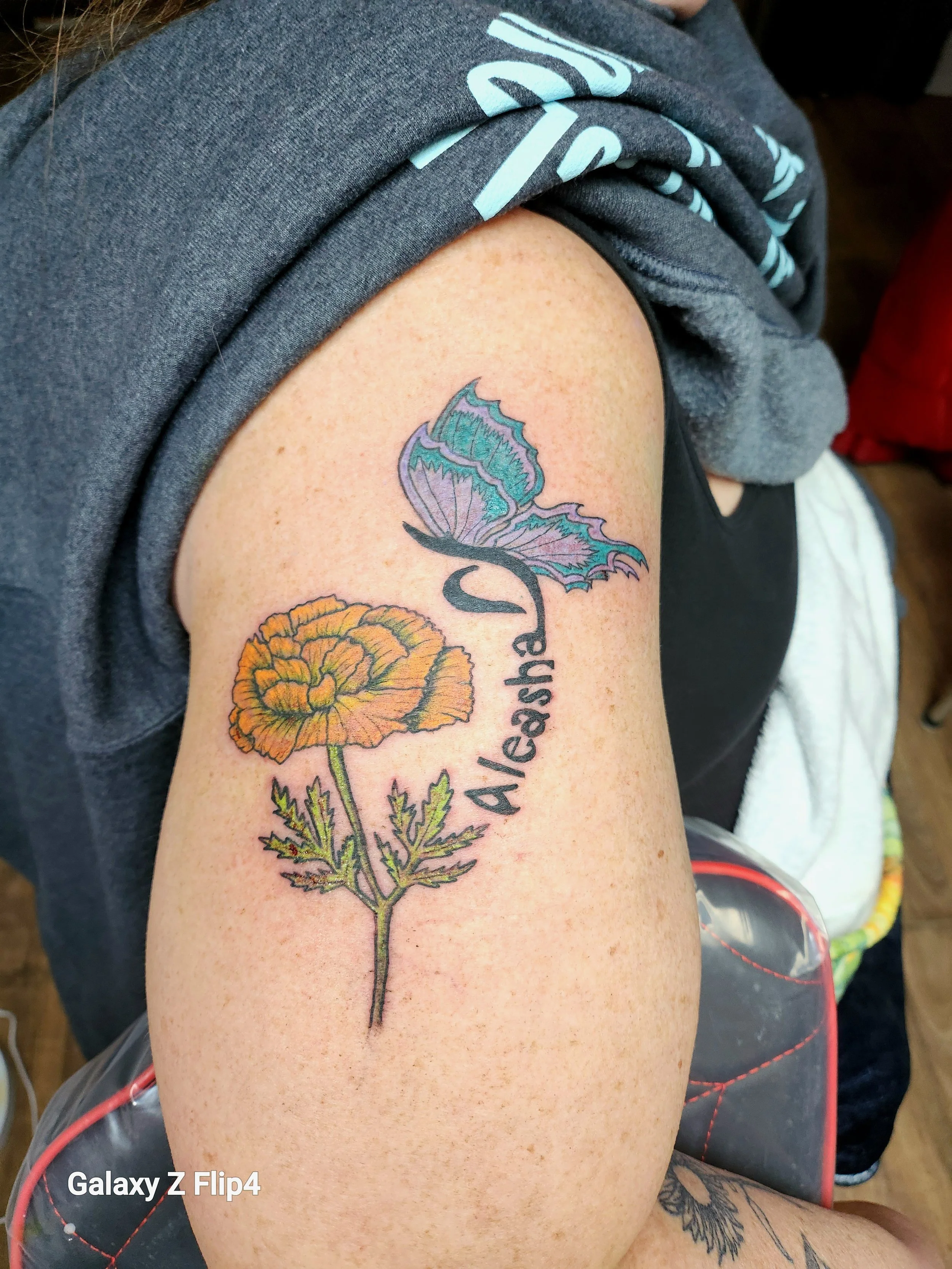 Tattoo of a yellow and orange sunflower with a teal and purple butterfly flying above it with the name 'Aleasha' underneath the butterfly