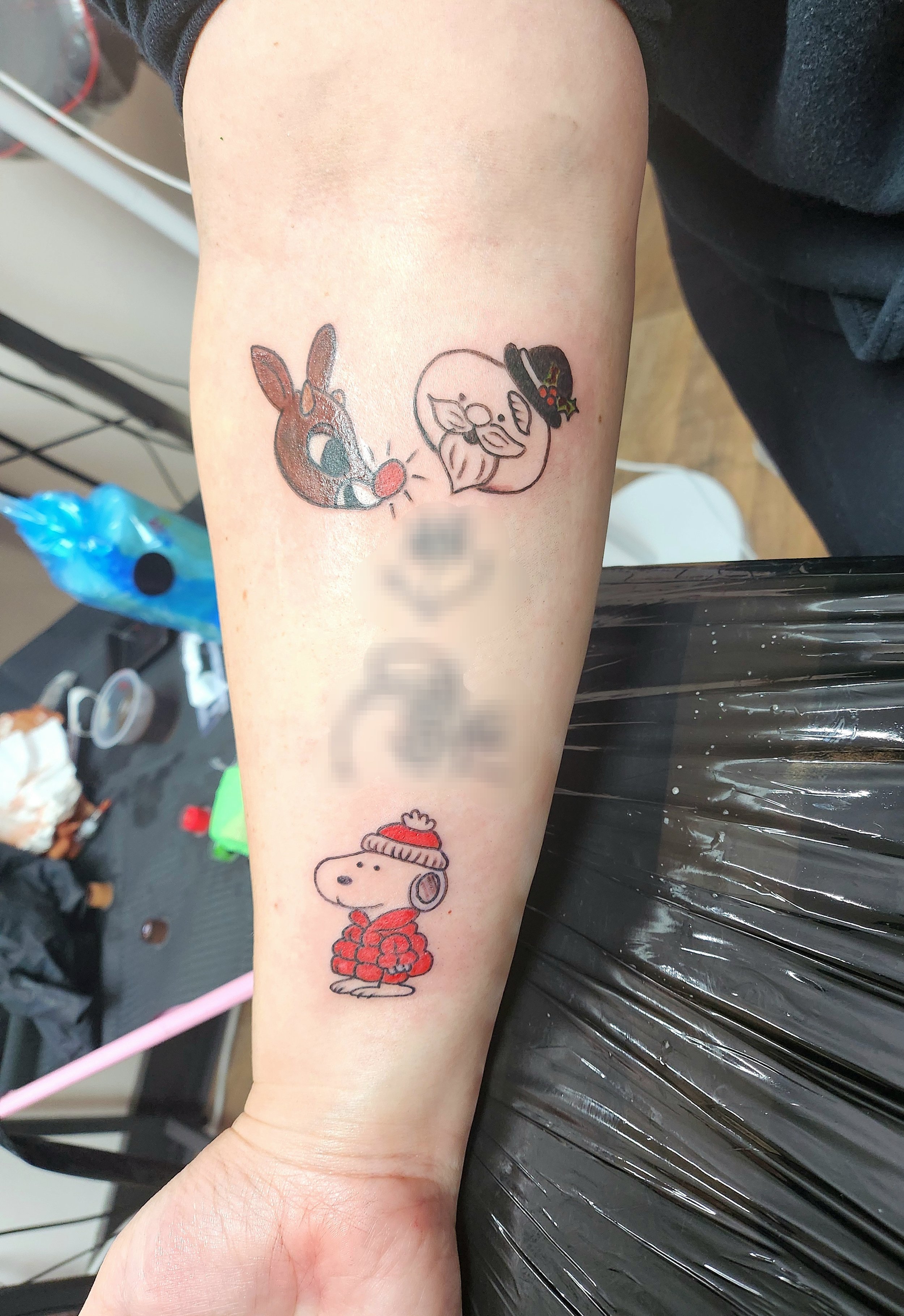 Three tattoos, all of which have a Christmas theme to them. The first one shows Rudolf the Red Nosed Reindeer, the second one is of the snowman host from Rudolf the movie, and the third is of Snoopy wearing a puffy red winter coat and red hat.