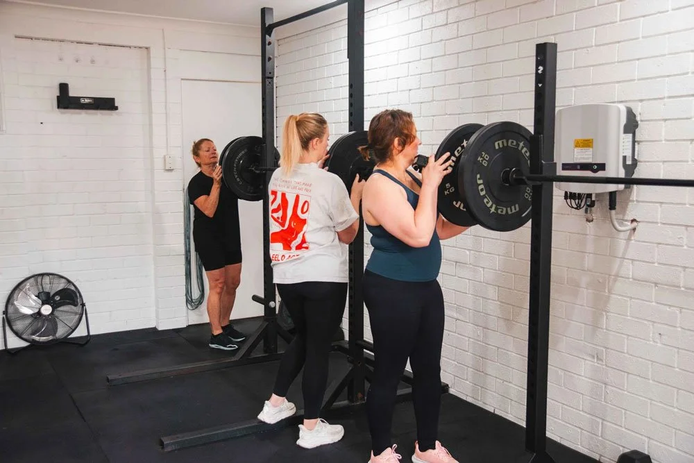 Three women in gym attire lifting barbells in a weightlifting room with white walls and black gym equipment.