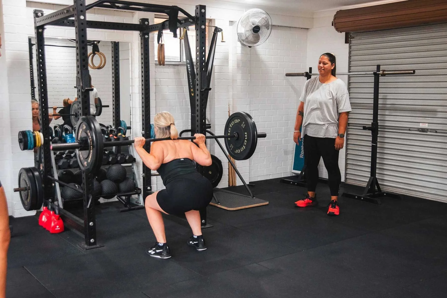Strength Training Classes for Women on the Central Coast:: A woman in black workout clothes squats with a barbell with weights on her shoulders in a gym, while a trainer observes her.