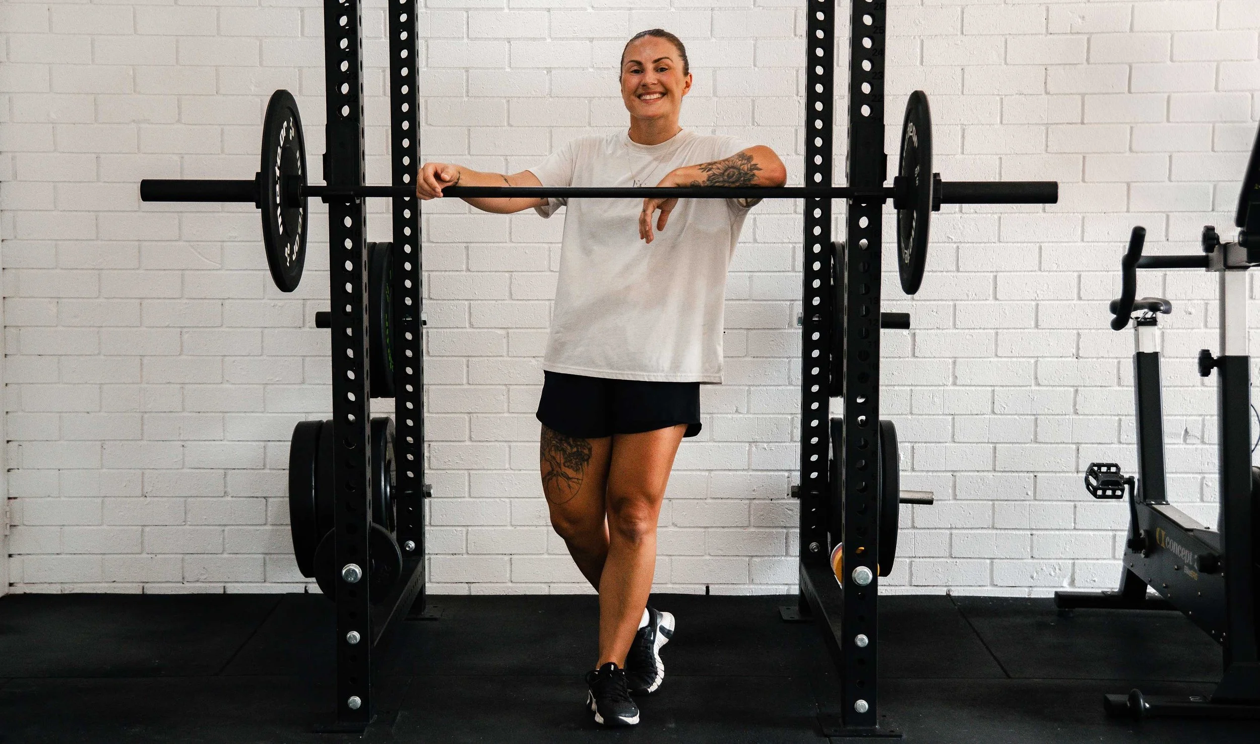 A woman smiling in a gym standing next to a power rack with a barbell, wearing a white t-shirt, black shorts, and athletic shoes.