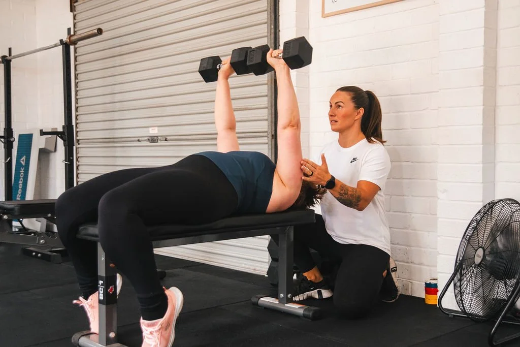 A woman assisting a person with a weightlifting exercise on a bench, inside a gym with a white brick wall and a large industrial fan on the floor.