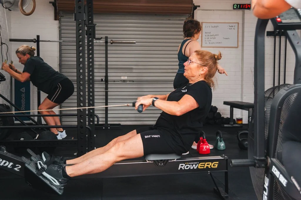 An woman in black workout clothes is doing rowing exercises on a rowing machine in a gym, with two kettlebells on the floor nearby and other people exercising in the background.