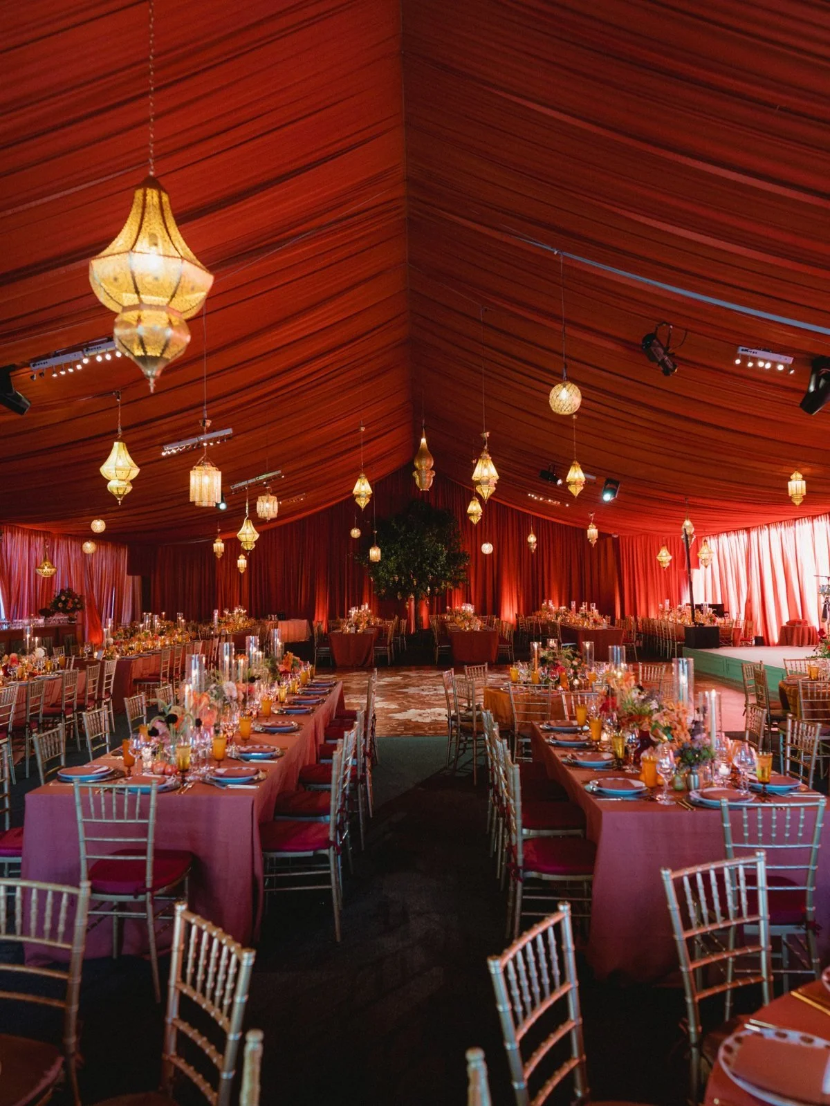 Moroccan-inspired wedding reception beneath a red draped tent with hanging lanterns and banquet tables