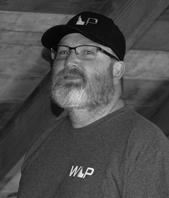 Black and white photo of a bearded man wearing glasses, a hat, and a t-shirt with "WLP" logo, looking towards the camera.
