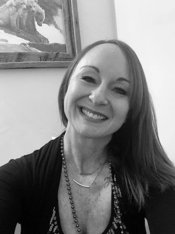 Smiling woman posing for a selfie with long hair, wearing a necklace, and seated indoors. Background features a framed bear painting.