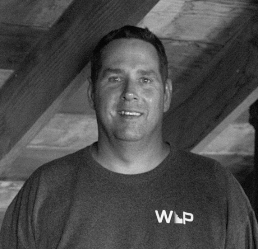 Man smiling in front of wood beams, wearing a shirt with "WLP" printed on it.