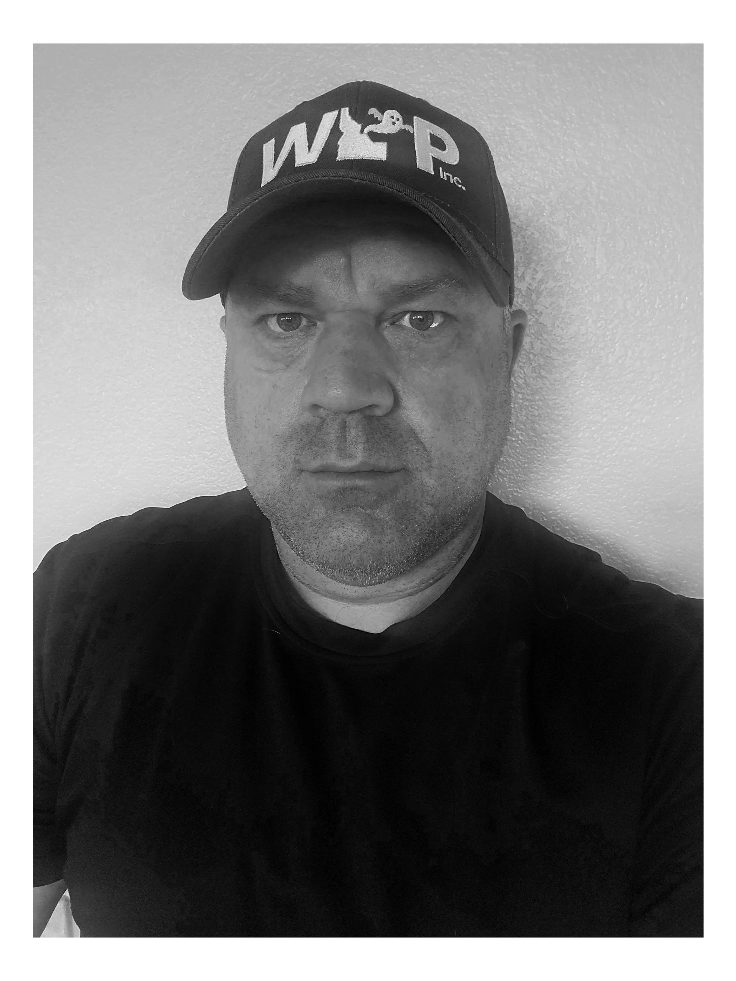 Black and white photo of a man wearing a cap with W2P Inc. logo, looking directly at the camera, against a plain wall.