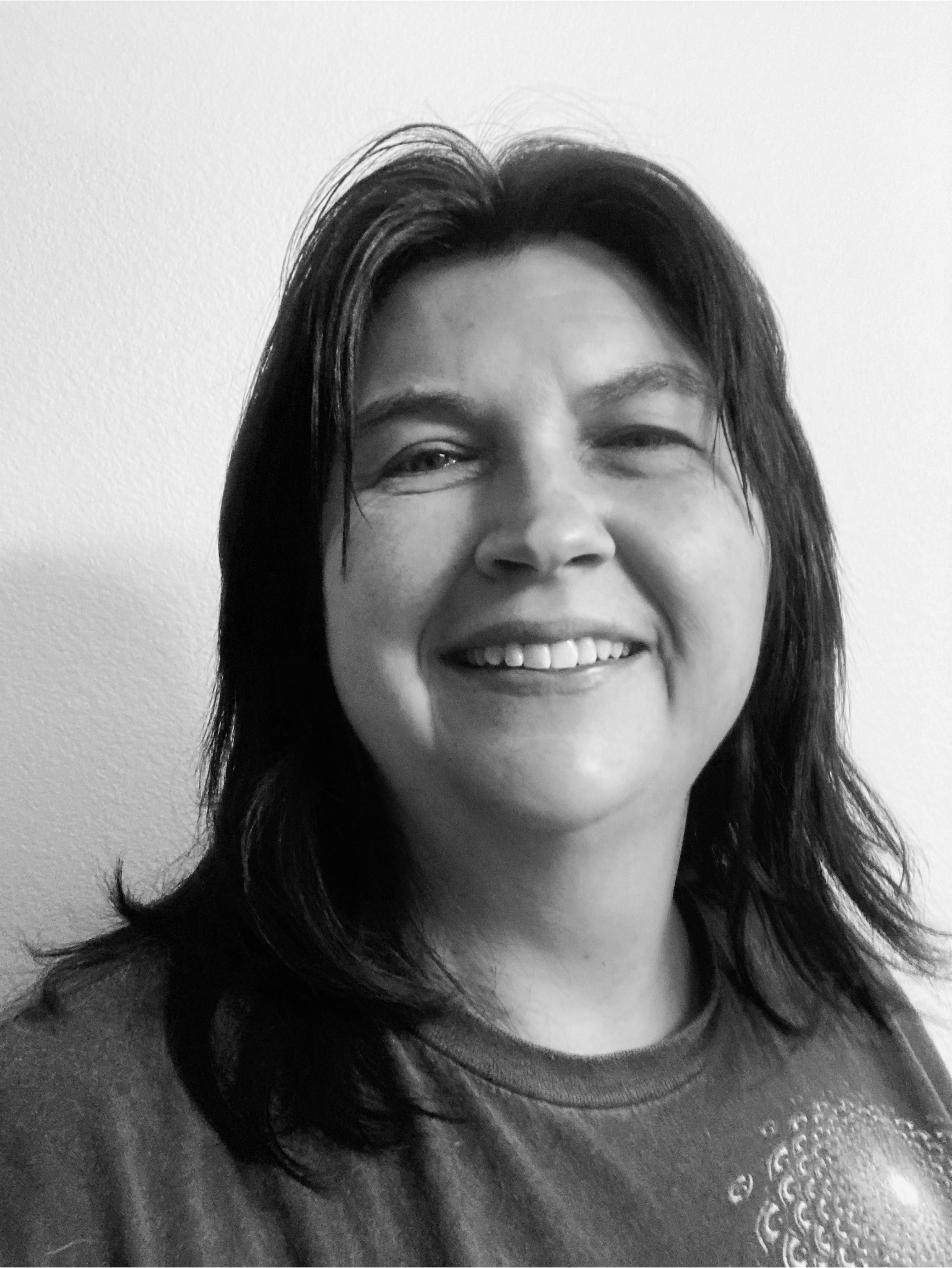 Black and white photo of a woman smiling, with shoulder-length dark hair and wearing a sweatshirt, against a plain wall.