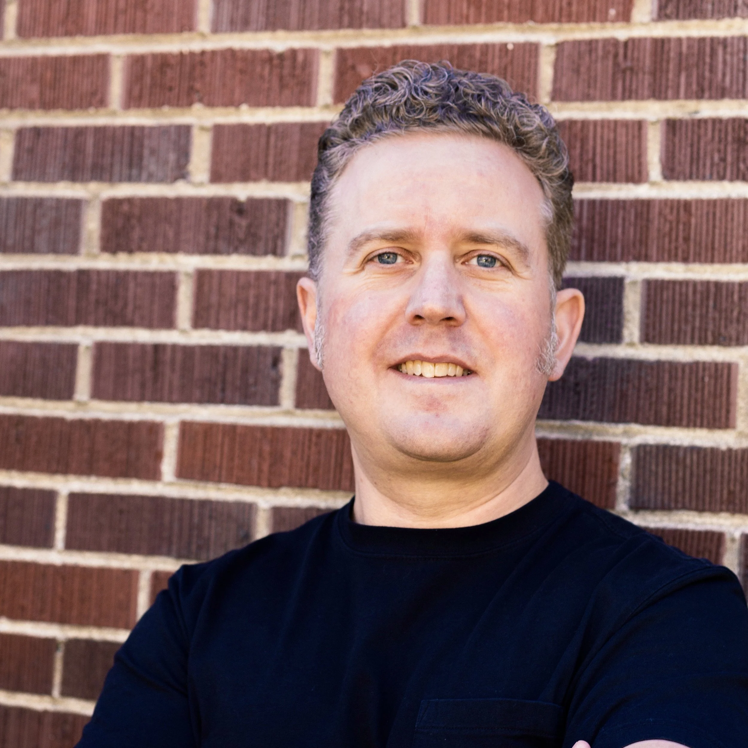 A man with curly hair and blue eyes standing in front of a brick wall, wearing a black shirt and smiling.