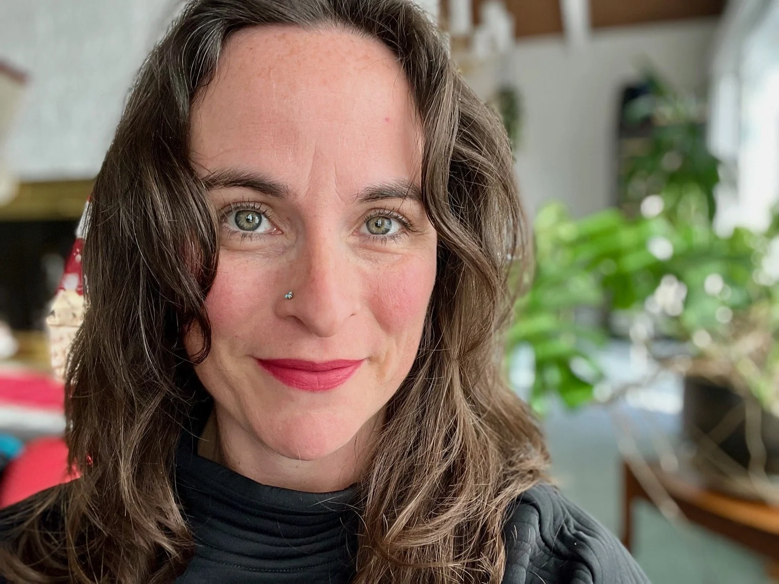 Close-up of a woman with wavy brown hair, gray eyes, and a small nose piercing, smiling indoors with green plants and sunlight in the background.
