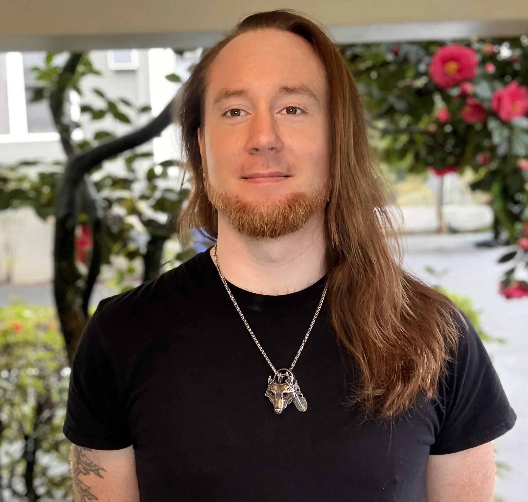 Portrait of a young man with long red hair, a beard, wearing a black t-shirt and a silver wolf head necklace, standing outdoors with trees and pink flowers in the background.