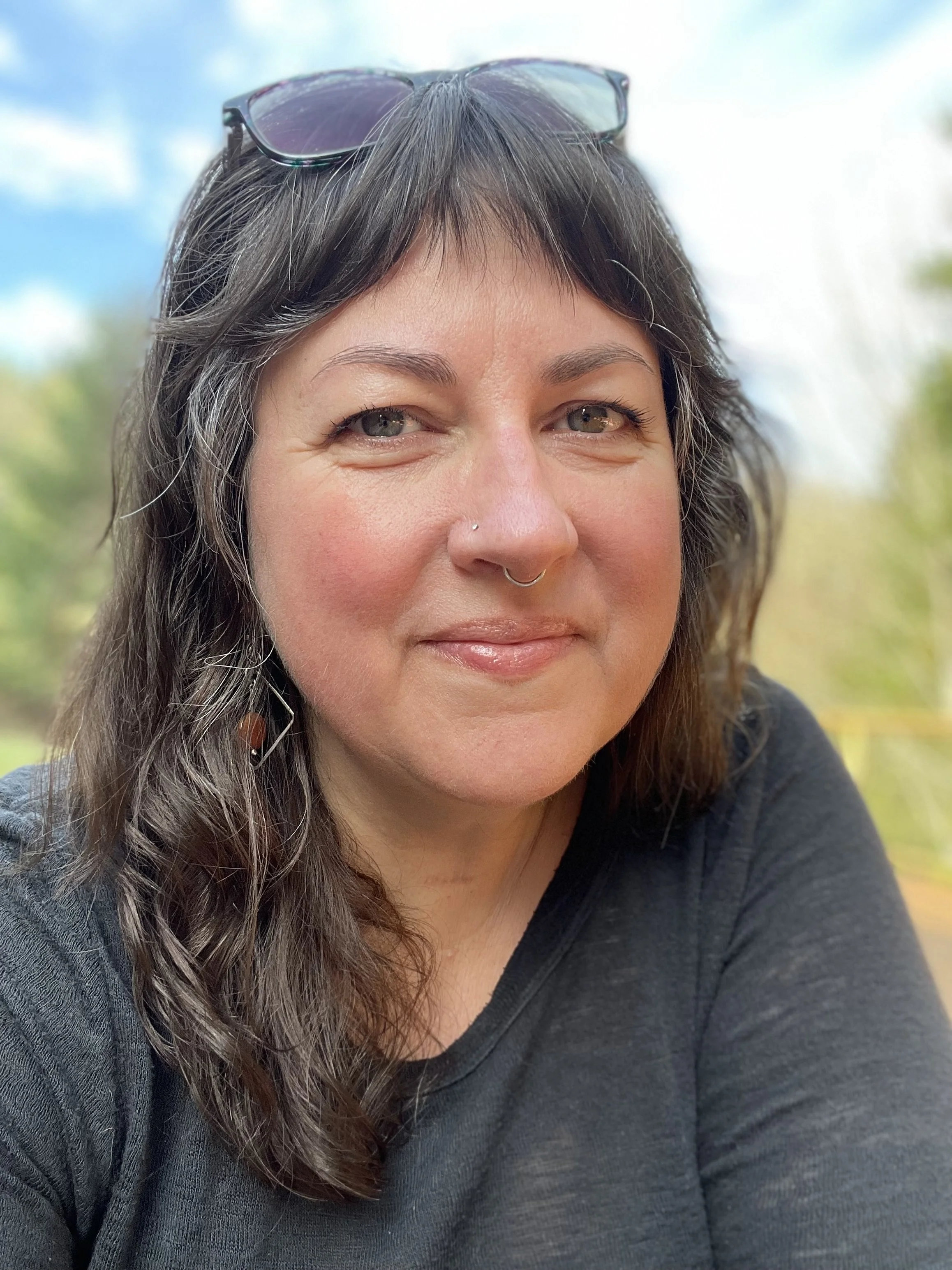 A woman with dark brown hair, sunglasses on her head, and a nose piercing, smiling outdoors on a partly cloudy day.