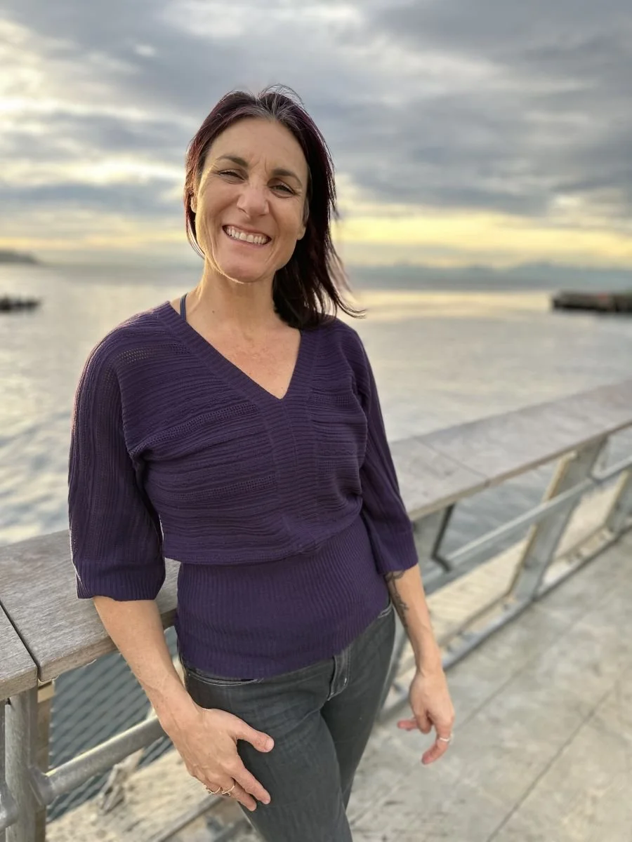 A woman with dark hair and fair skin smiling and leaning on a wooden railing by a body of water during sunset.