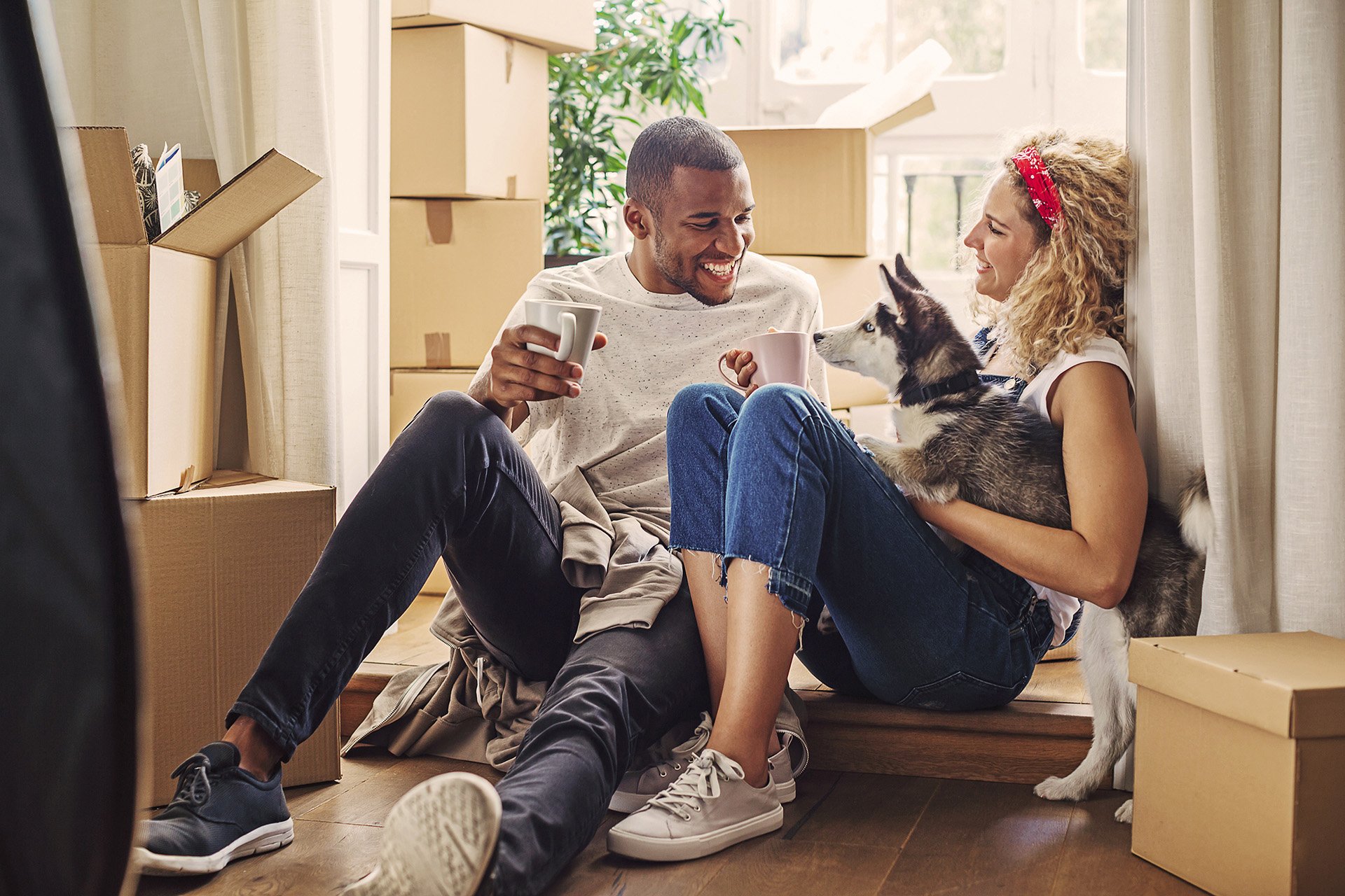 Couple sitting on floor with coffee mugs, surrounded by moving boxes, holding a puppy.