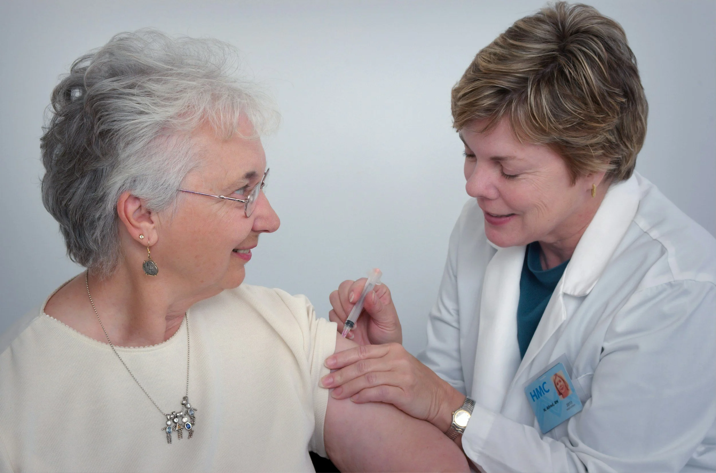 A healthcare professional administers a vaccine to an elderly woman in a clinical setting. The woman is smiling and wearing glasses and jewelry, while the healthcare provider is focused on the injection, dressed in a white coat with a visible name badge.