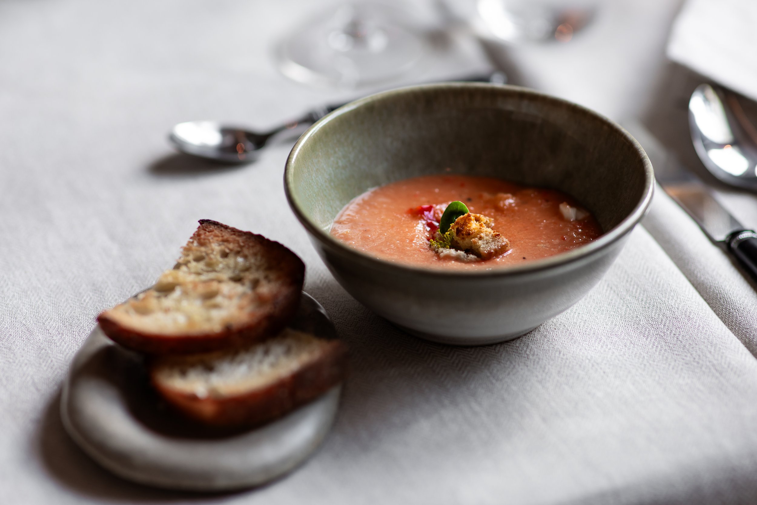 En skål med tomatsoppa toppad med krutonger och örter, bredvid en tallrik med skivor av rostat bröd.