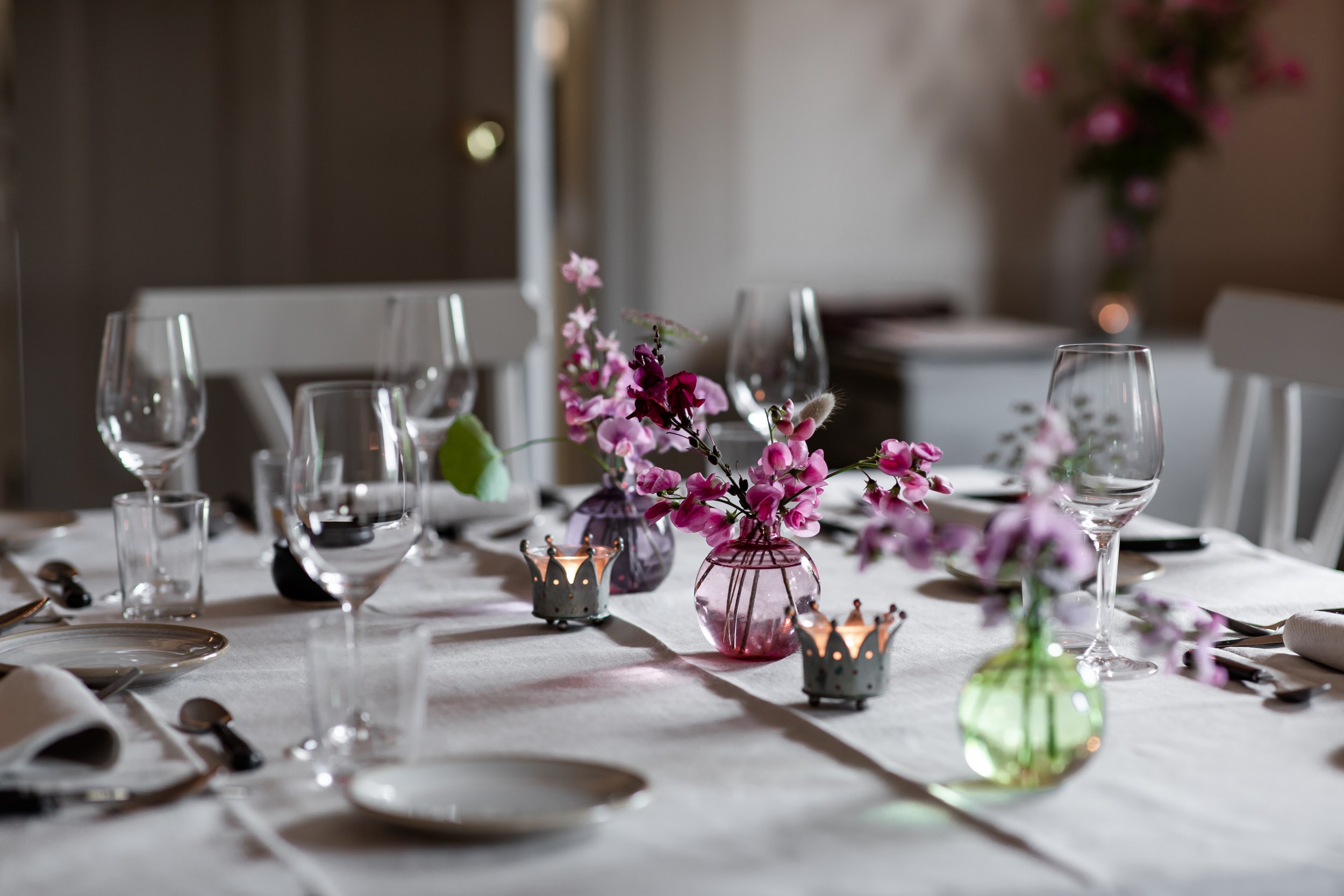 Ett vackert dukat bord med vita dukar, vinglas, tallrikar och bestick. Små vaser med rosa blommor och metalliskt formade ljushållare pryder bordet.
