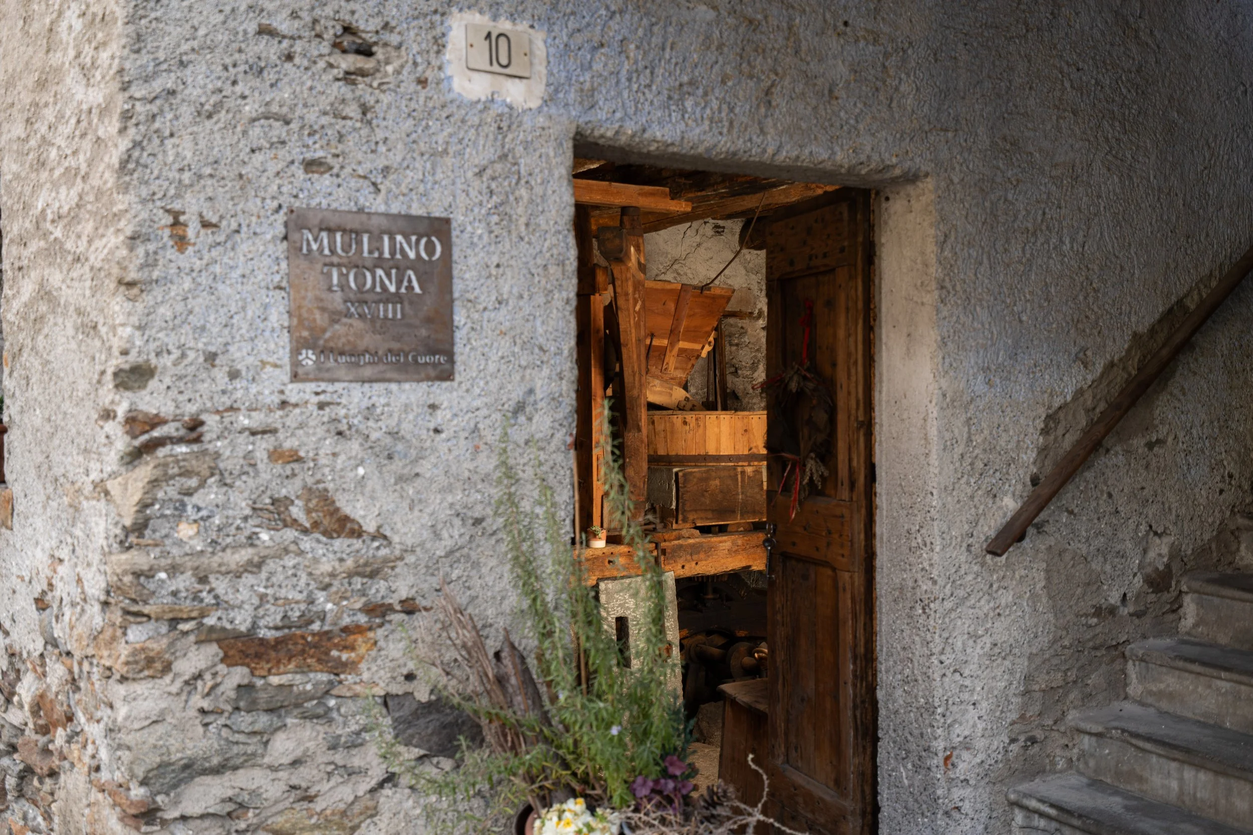 The image shows a rustic stone wall with a plaque and a doorway leading into a wooden interior space. The plaque reads 'MULINO TONA XVII'. The doorway opens into what appears to be an old mill or workshop with wooden beams and equipment inside.