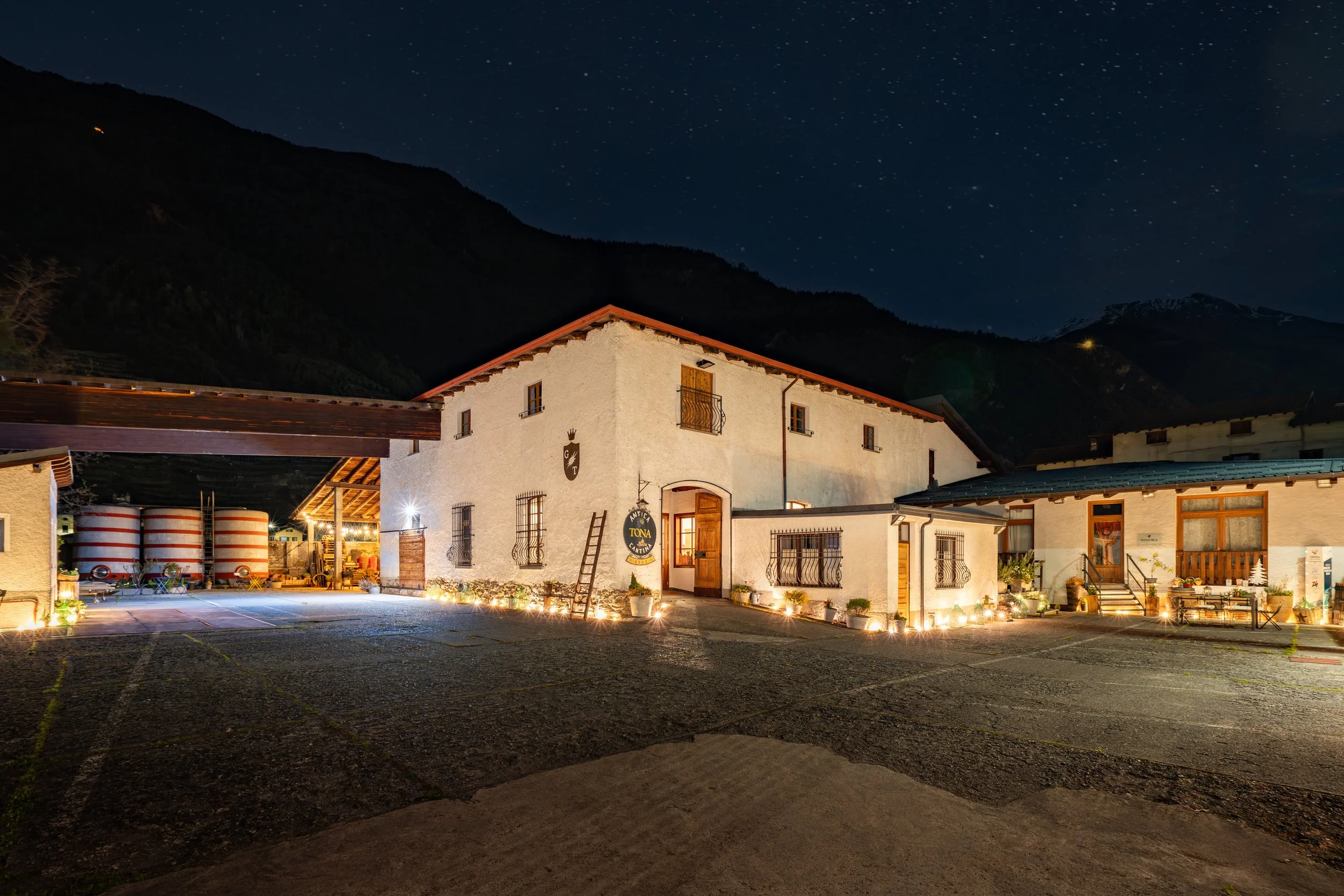 Nighttime scene of a white building with a red roof, illuminated by exterior lights, surrounded by a paved area, with mountains and a star-filled sky in the background.