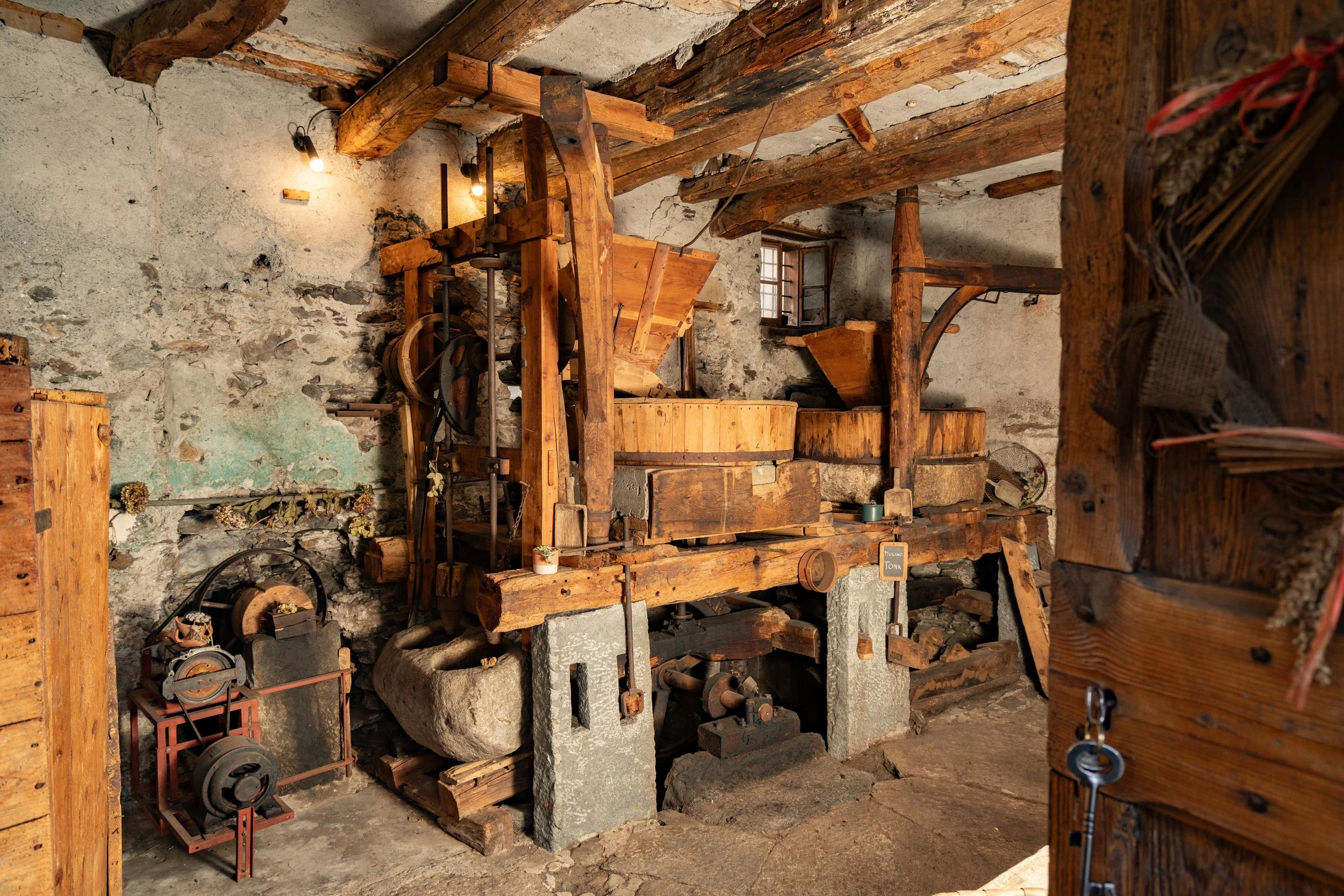 Interior of an old rustic workshop or mill with wooden beams, machinery, and concrete foundation. The space features vintage tools, a large stone, and some labeled equipment, illuminated by minimal lighting.