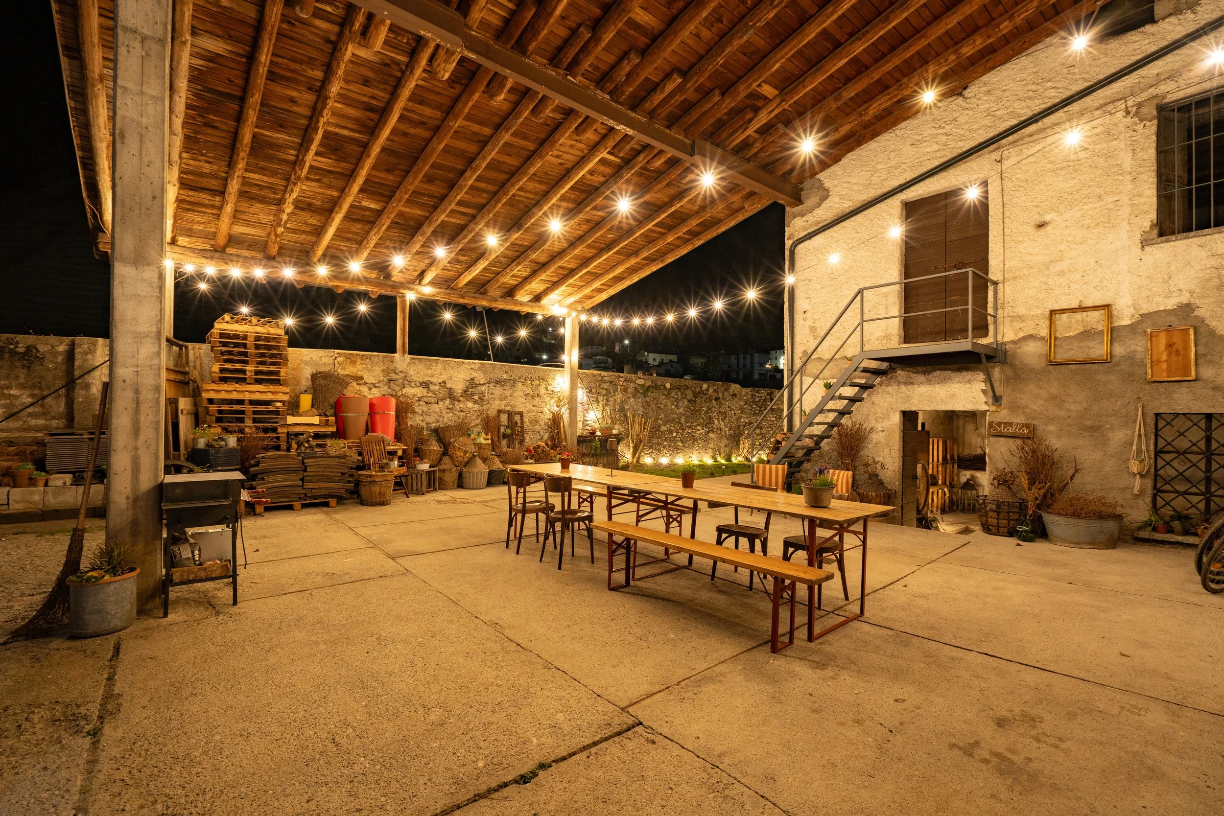 Night view of an outdoor courtyard with string lights, a wooden roof, a stone wall, metal staircase, and patio furniture, including tables and chairs.