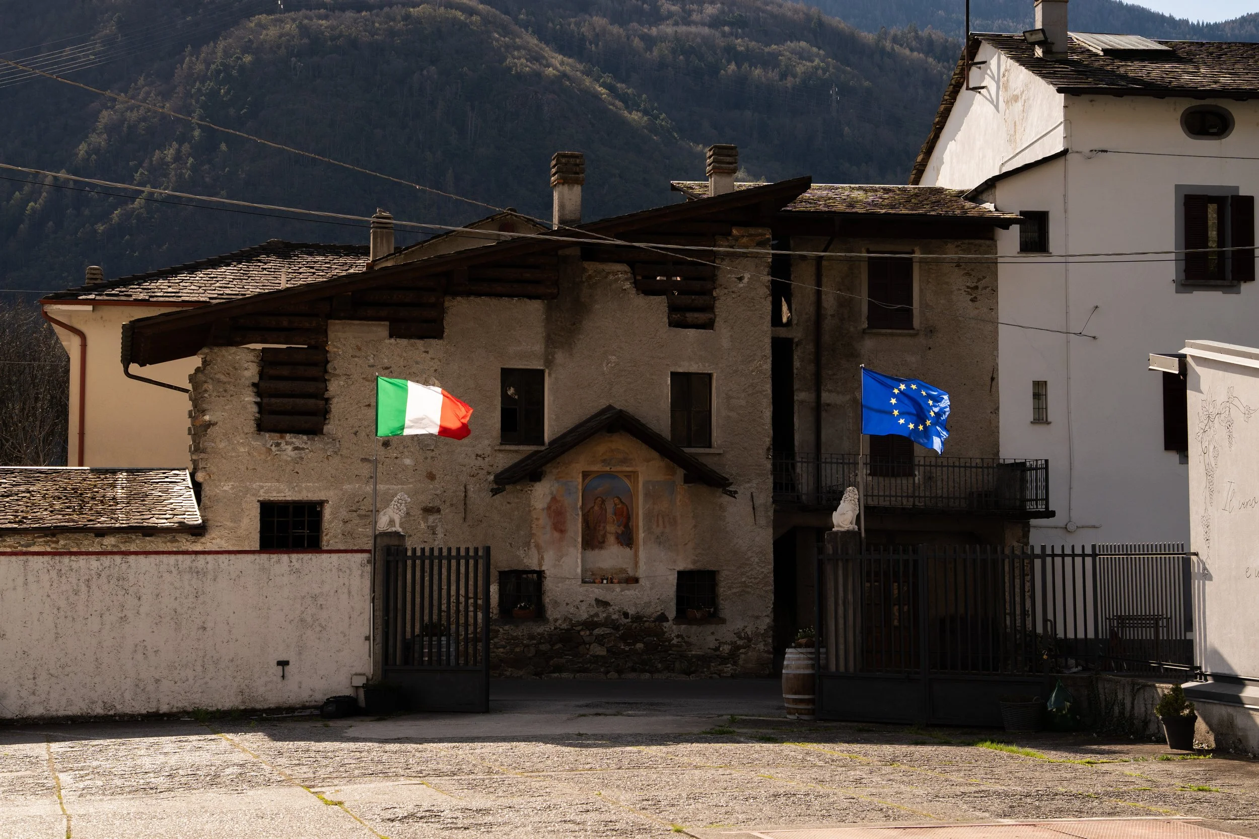 A small European village with old stone and stucco buildings, featuring two flags (Italy and European Union) mounted on poles, set against a mountainous backdrop with forested hills.
