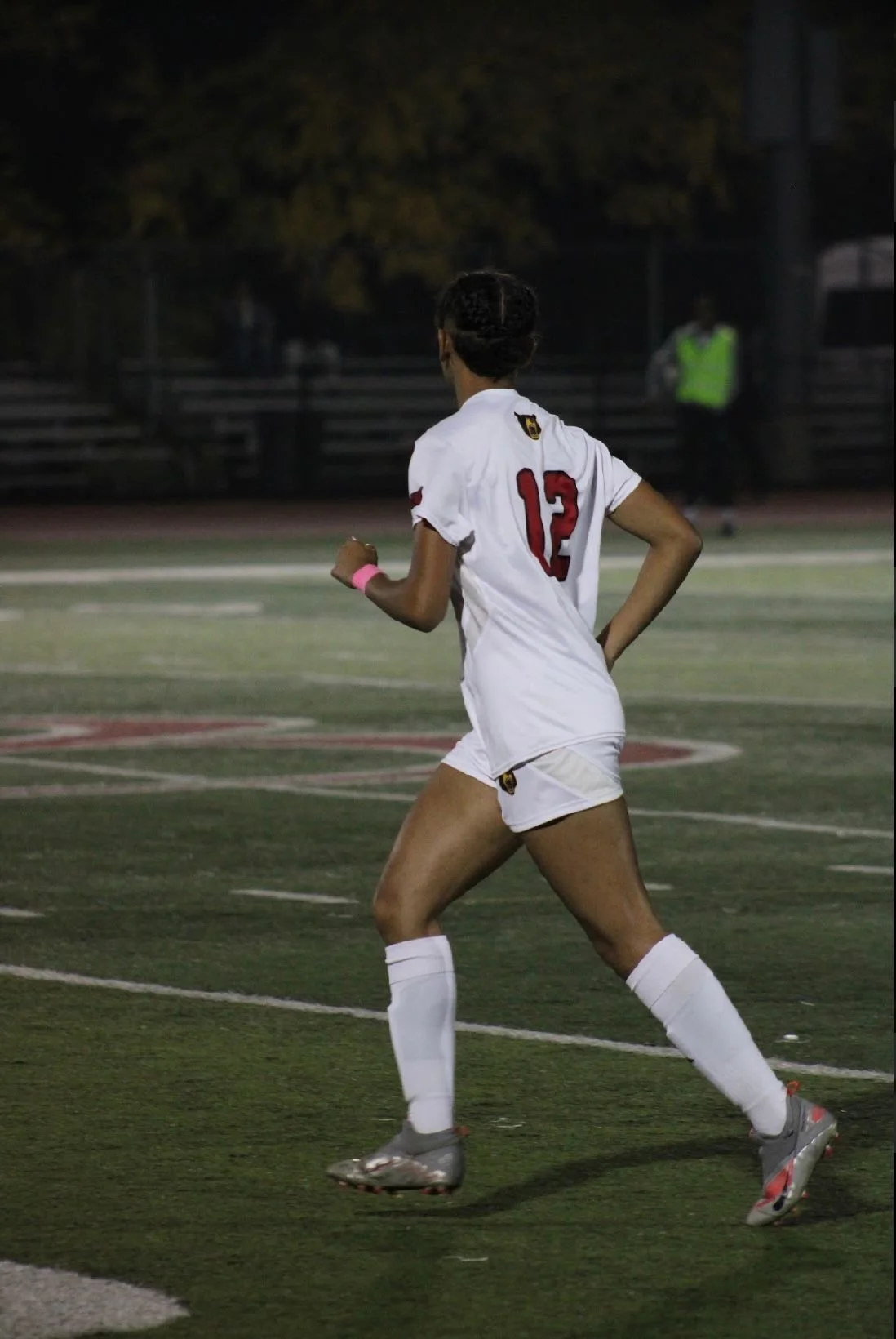 A female soccer player wearing a white uniform with the number 13 on her back, running on a soccer field at night.