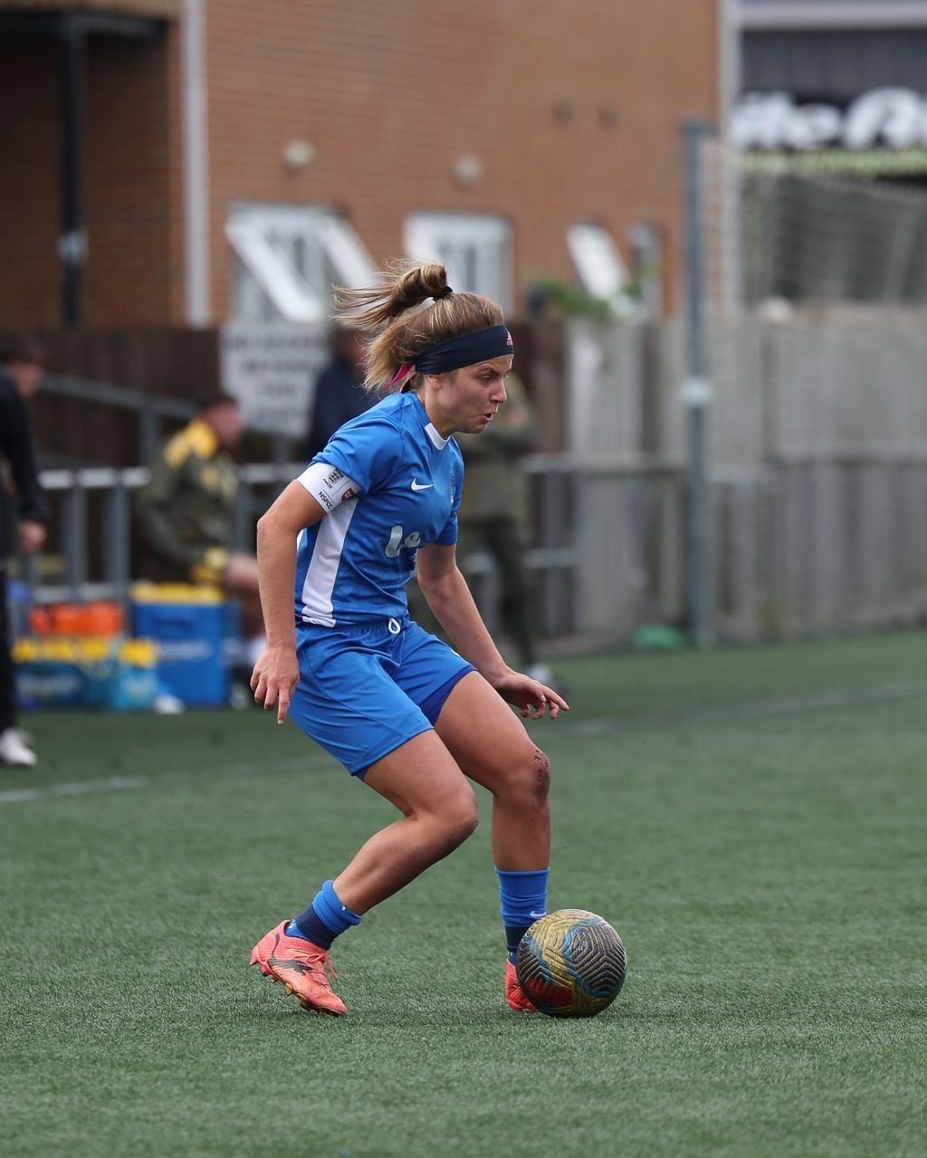 Female soccer player in blue uniform and pink cleats preparing to kick a soccer ball on a field.
