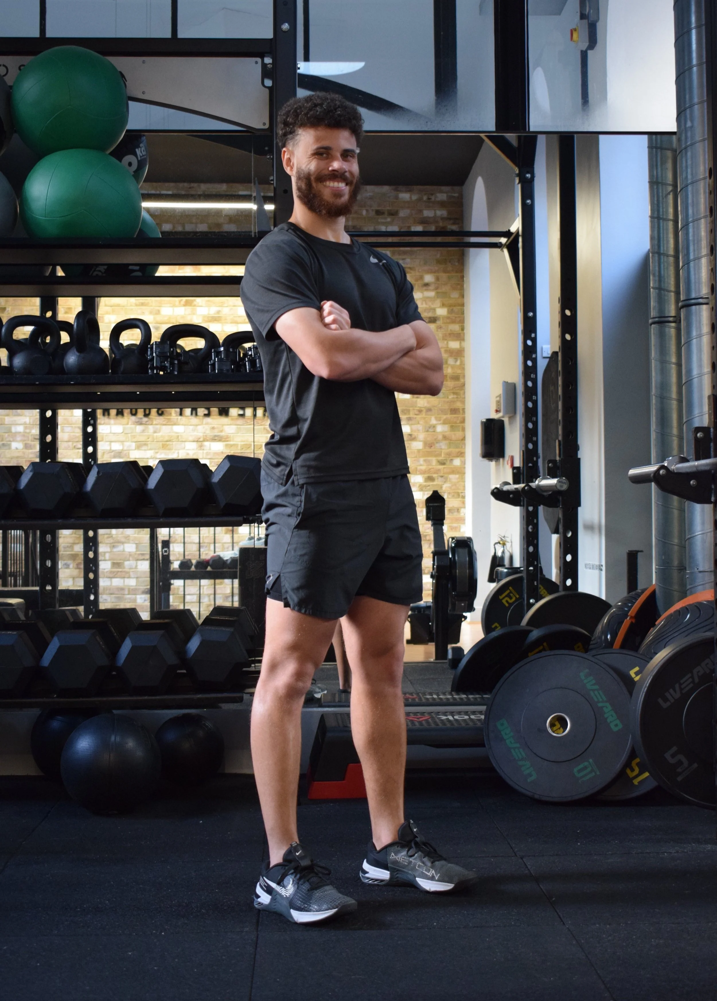 A smiling man standing with arms crossed in a gym, surrounded by exercise equipment including dumbbells, kettlebells, and weight plates.