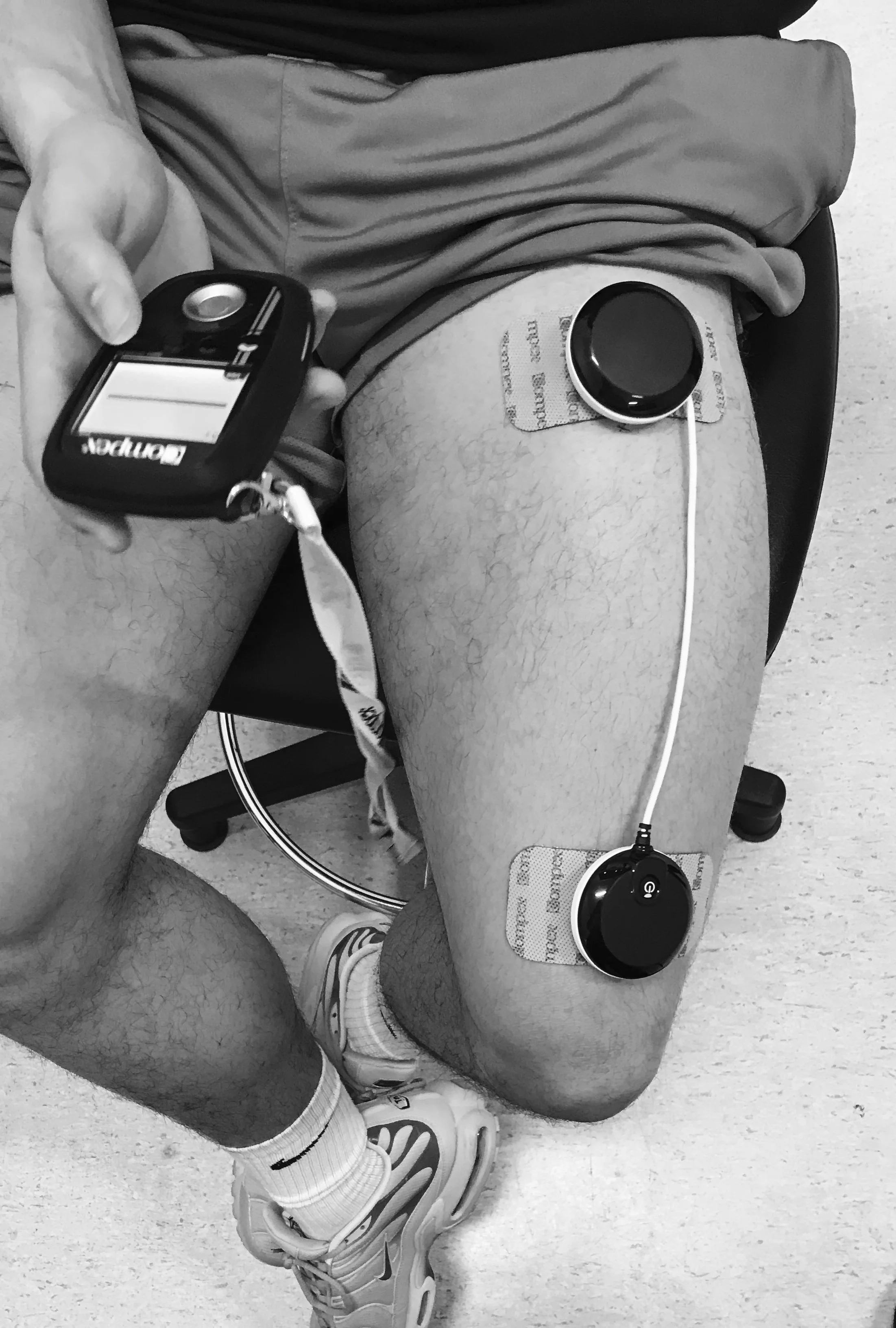 A person lying on a chair with a blood glucose meter in one hand and an insulin pump attached to their thigh with adhesive pads, wearing athletic shoes and socks.