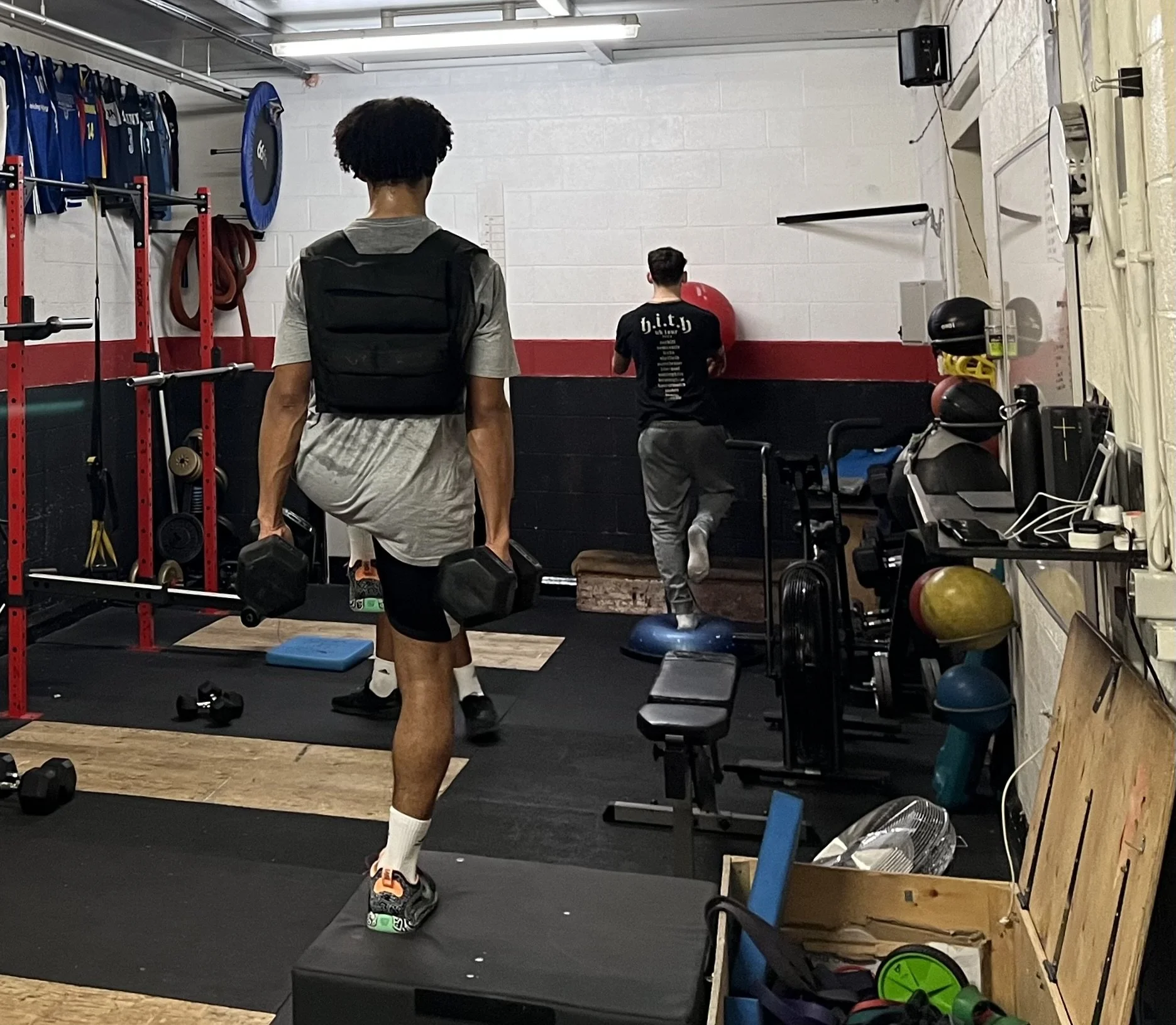 Two men exercising in a gym. One is walking on a step platform holding dumbbells, and the other is stretching against a wall with a medicine ball.