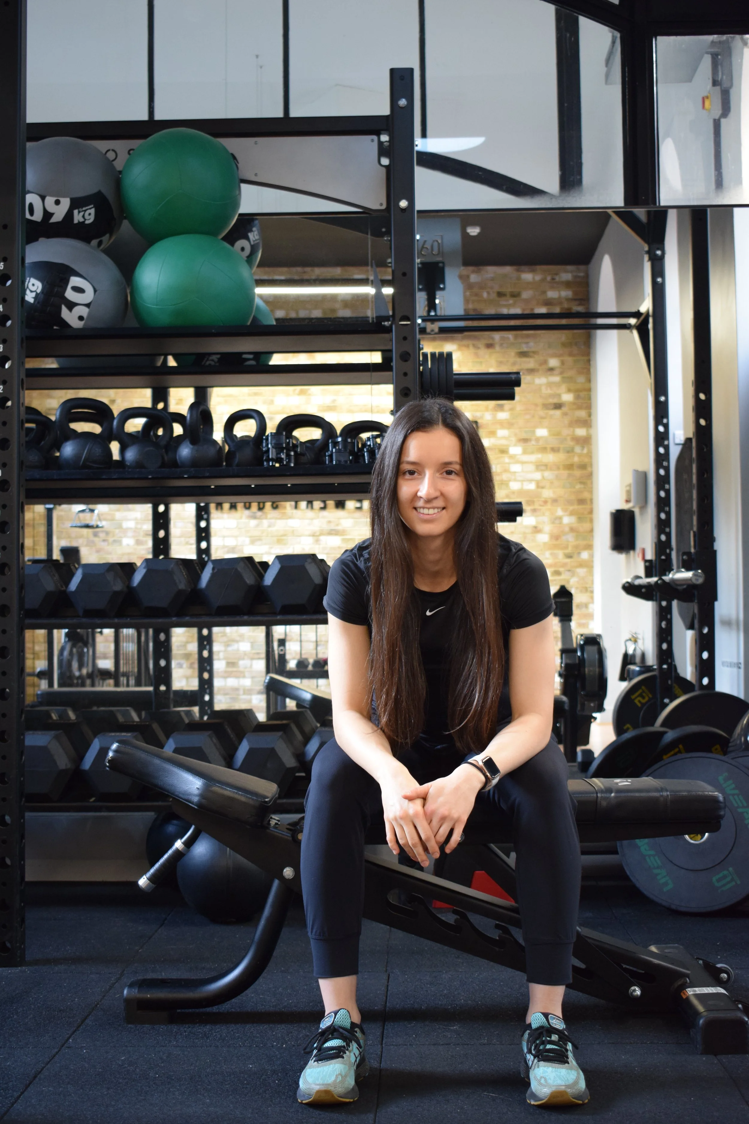 A woman sitting on a workout bench in a gym surrounded by kettlebells, dumbbells, and exercise balls.
