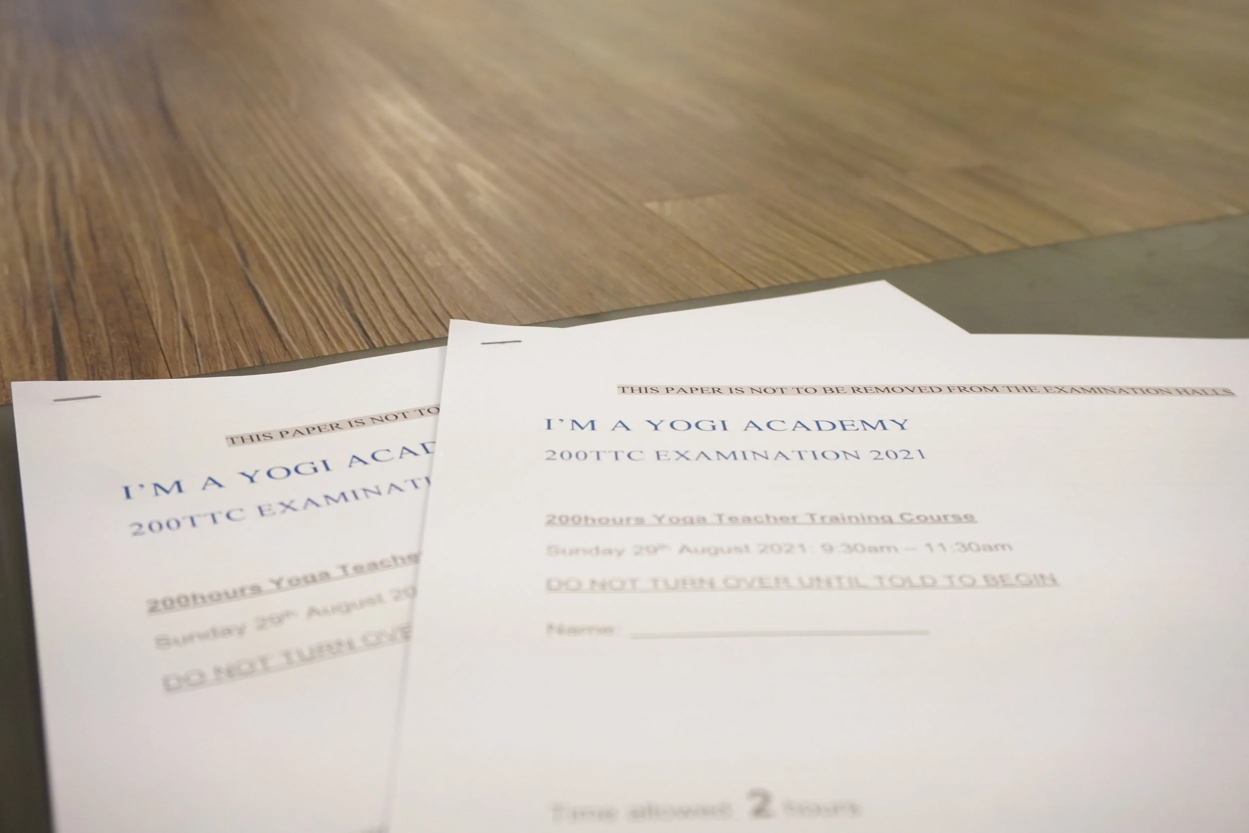 Two printed exam papers titled 'I'm a Yogi Academy 200 TTC Examination 2021' placed on a wooden surface.
