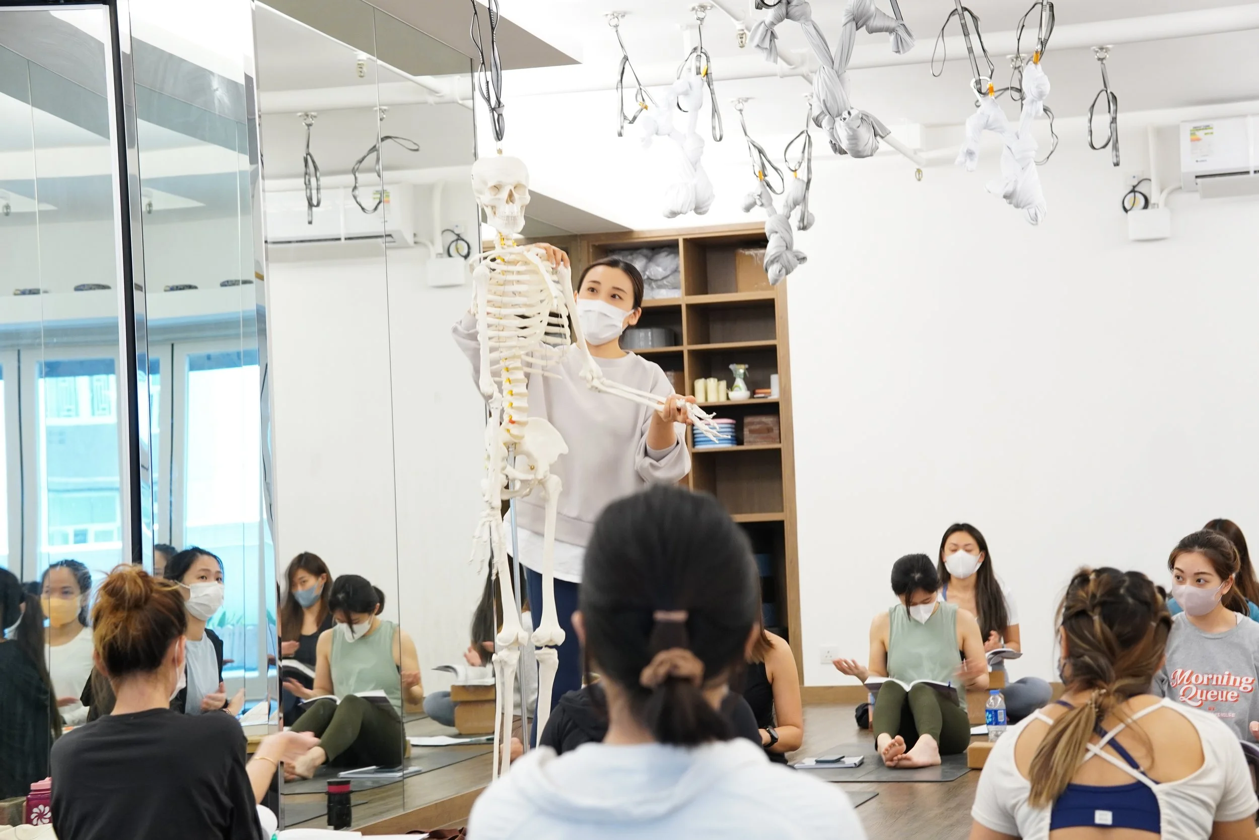 A woman giving a health or anatomy class using a human skeleton model to a group of students in a classroom. All individuals are wearing face masks, and there are hanging skeletons or cloth-covered objects suspended from the ceiling.