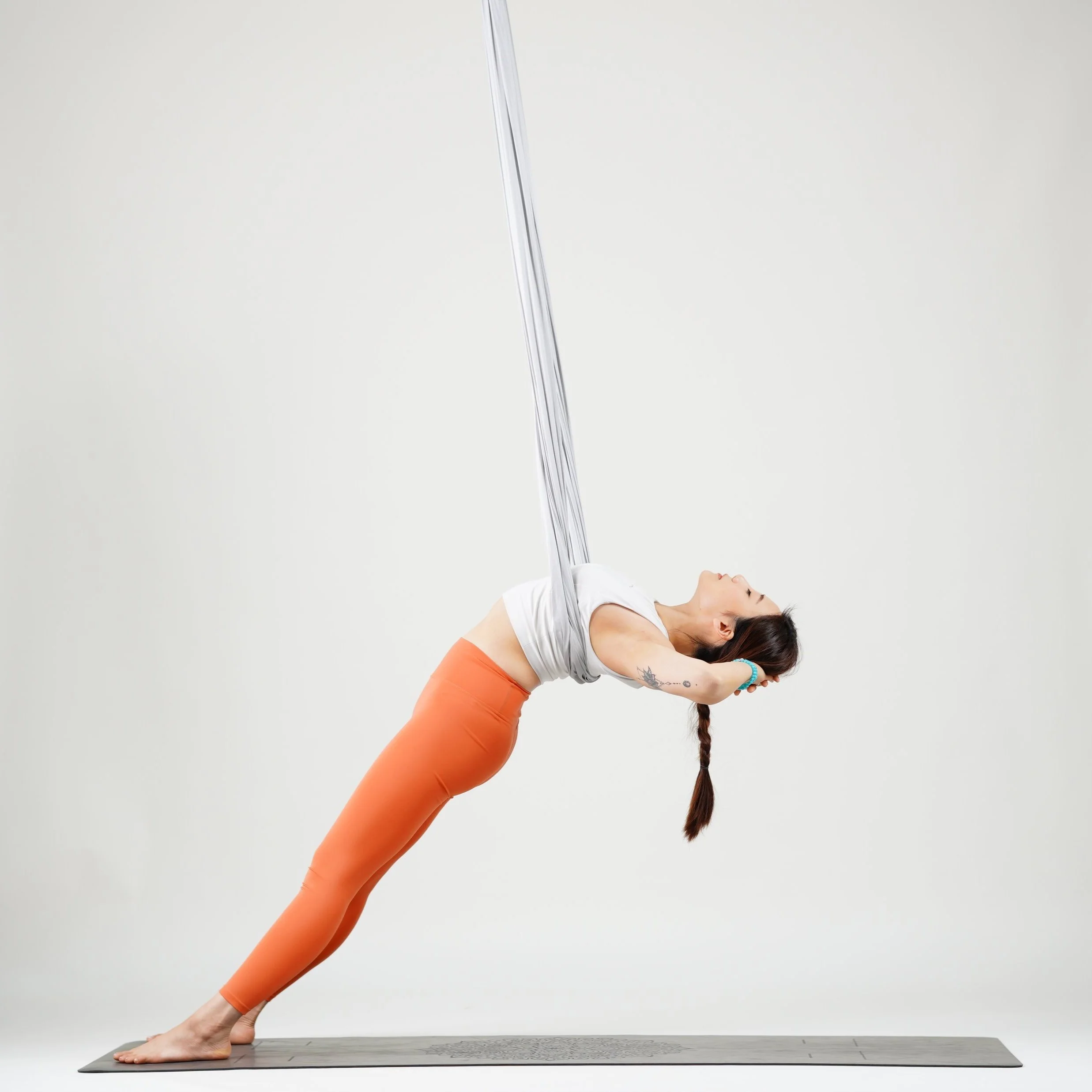 A woman practicing aerial yoga using silk hammocks in a studio with a plain white background. She is in a forward bend pose, with her hands clasped behind her neck, on a yoga mat.