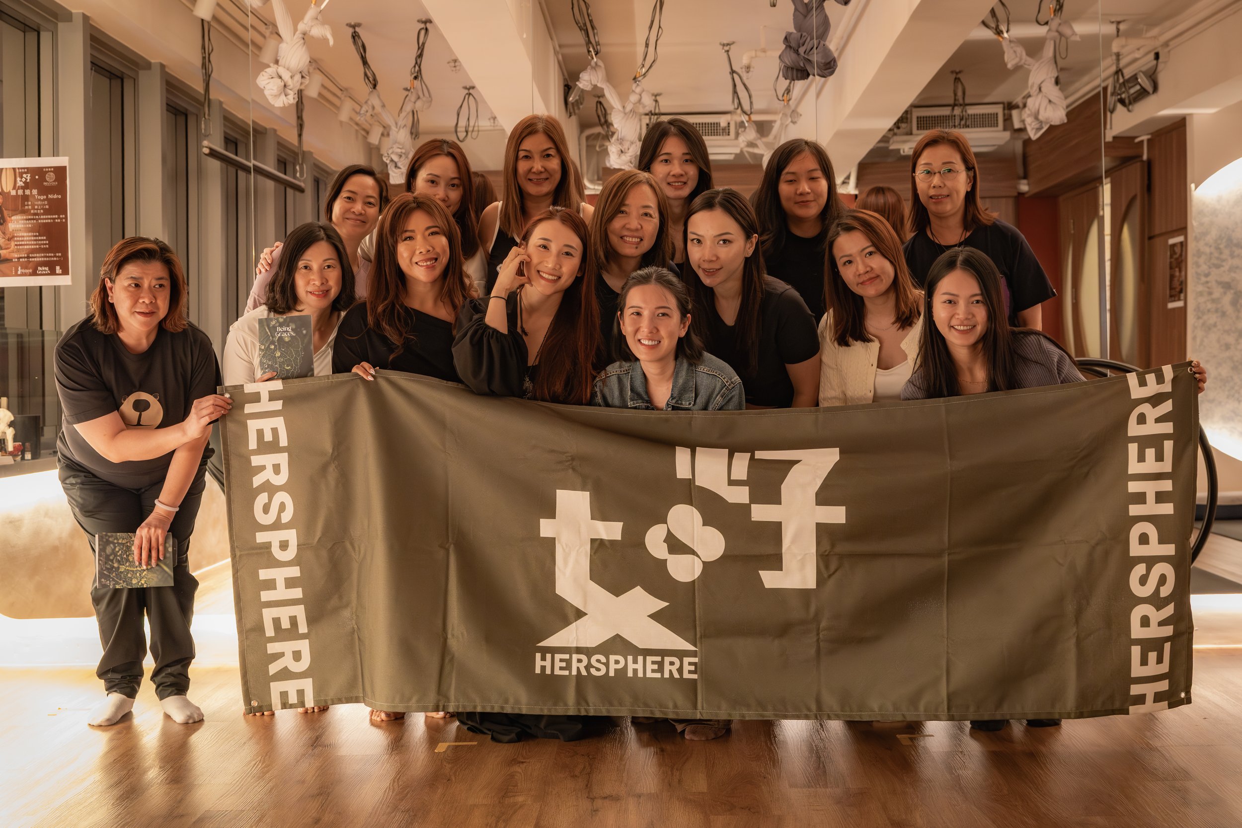 Corporate yoga class organized by IMAYOGI - A group of women posing together in a room, holding a large green banner with the words 'HERSHARE' and logos. They are smiling and standing close to each other, with some leaning on the banner.