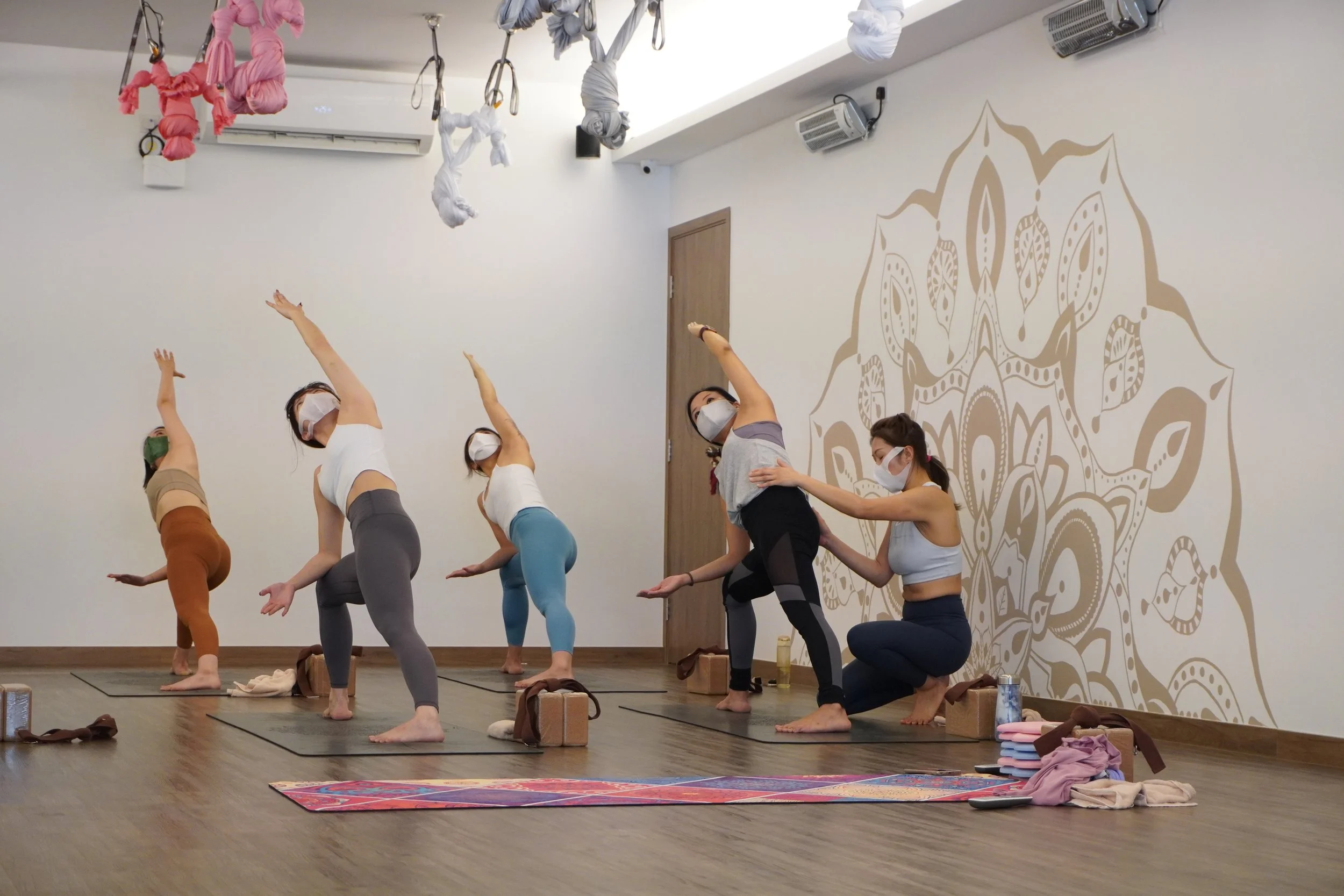 A group of five women practicing yoga in a studio, wearing masks, with mats and personal belongings nearby, and decorative aerial yoga hammocks hanging from the ceiling.