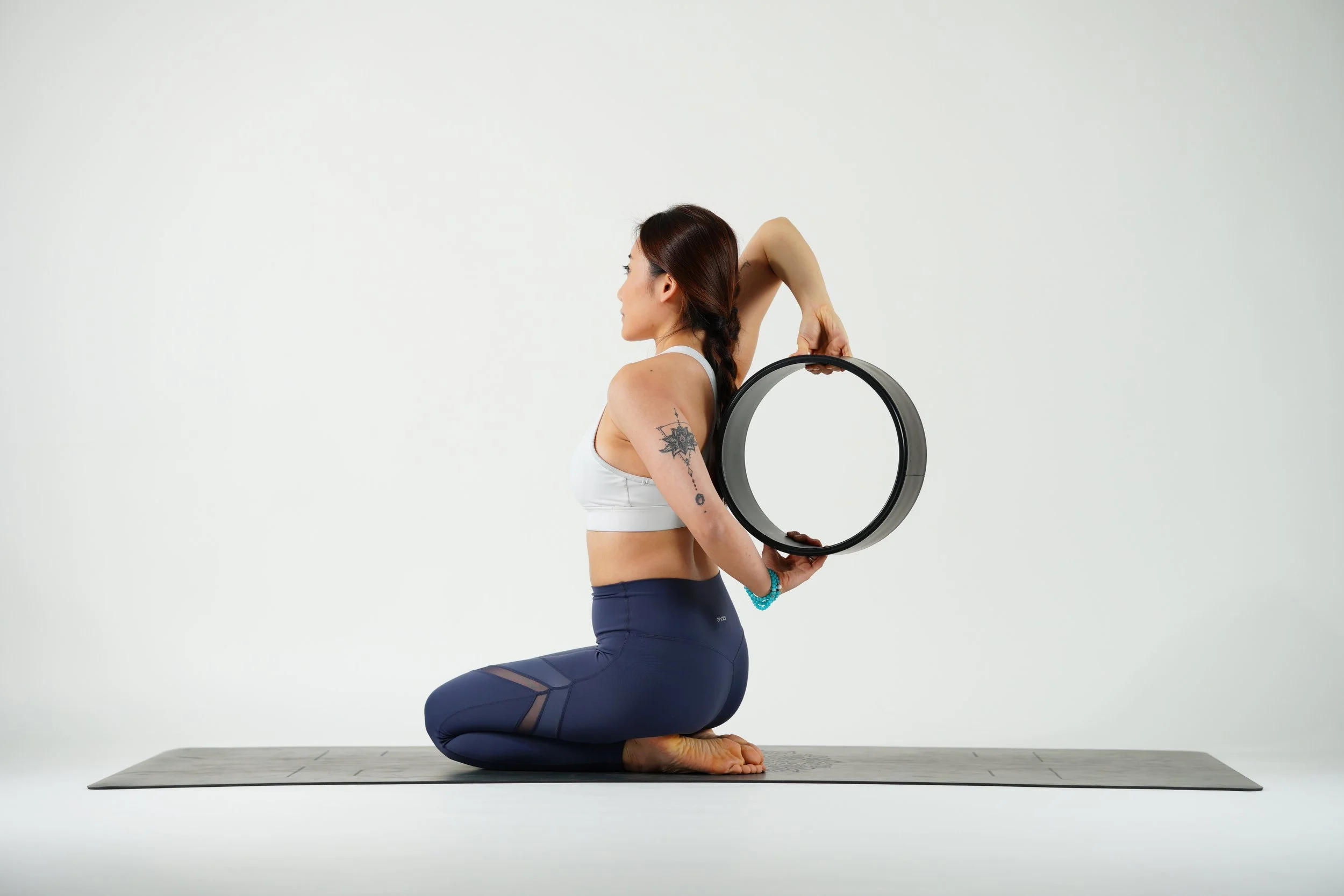 A woman practicing yoga kneeling on a yoga mat, holding a black yoga wheel behind her back against a plain white background.