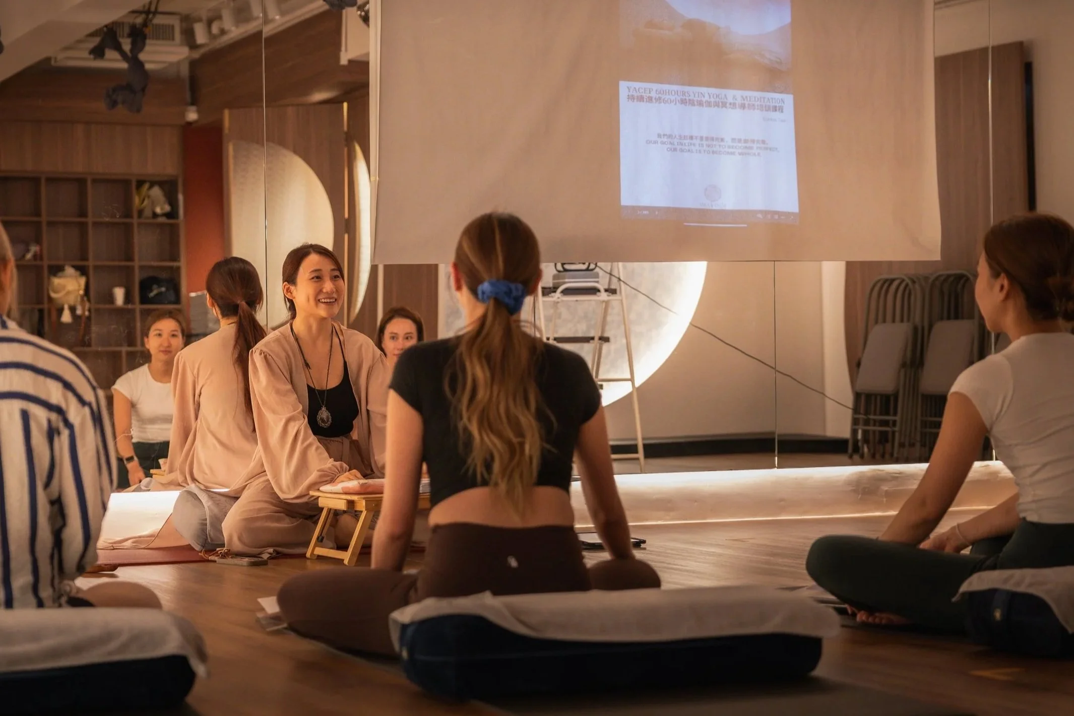 A group of women participating in a yoga, yin yoga, and meditation class, seated on cushions on the floor, facing a large screen with a presentation. The room has warm lighting and wooden decor.