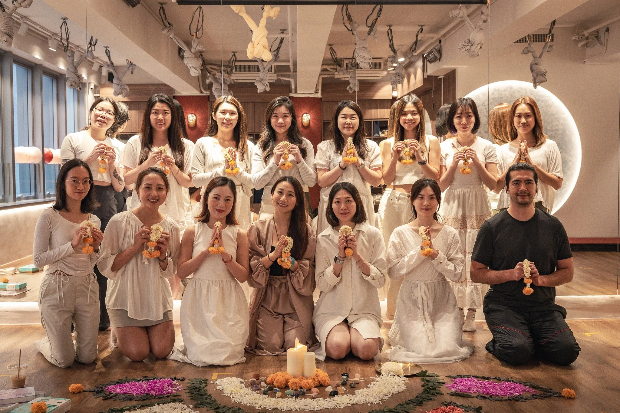 Group of women posing in a cultural celebration, dressed in white, holding flower garlands, with marigolds and offerings on the floor. They are standing and kneeling on a wooden floor in front of a wall with a decorative background.