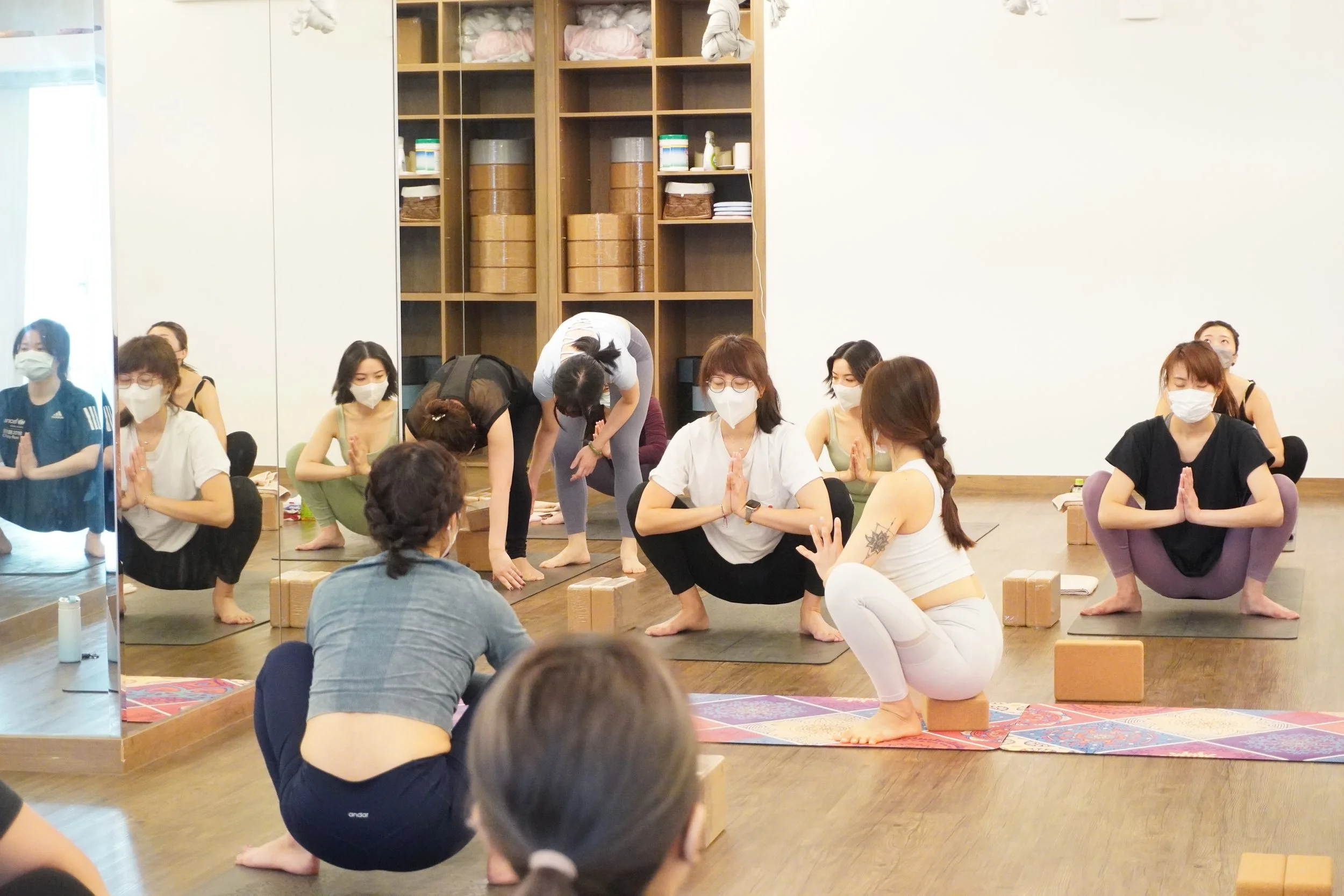 Women participating in a yoga class in a studio, all wearing masks, some in squatting positions and others doing different yoga poses, with blocks and mats on the floor.