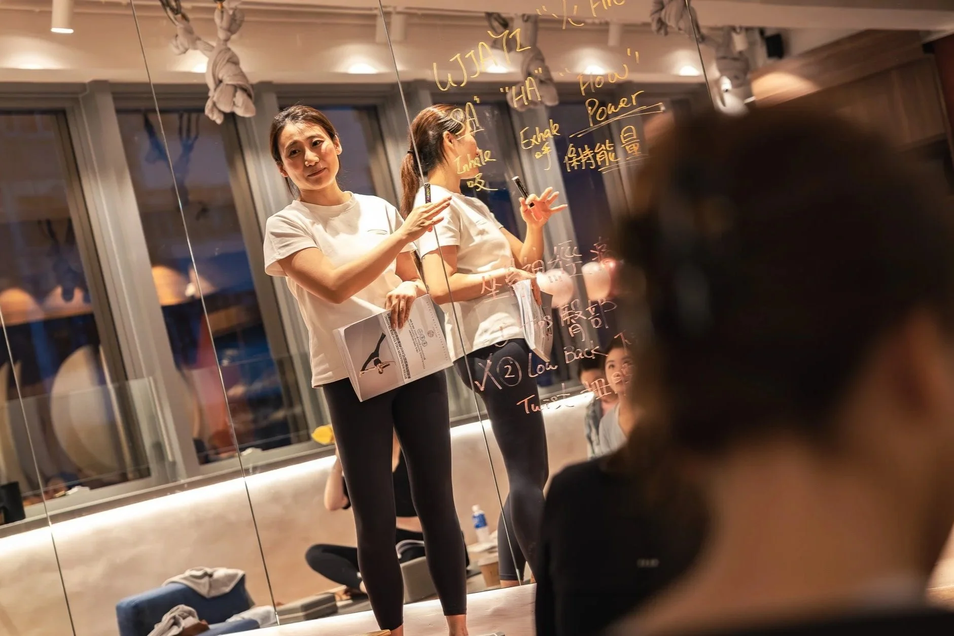 Women participating in a yoga or meditation class, sitting on yoga mats with some taking notes, while an instructor writes on a whiteboard.