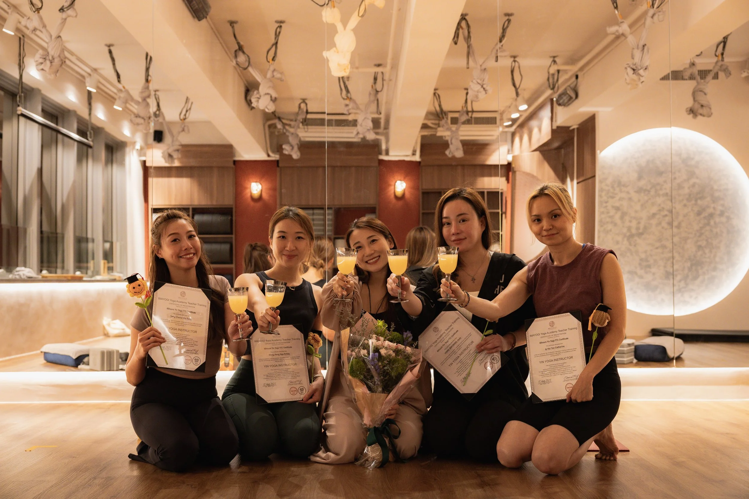 Five women kneeling on the floor inside a dance studio, holding certificates and glasses of orange juice, smiling for the camera. One woman in the middle holds a bouquet of flowers. The studio has a reflective wall, wooden floors, and decorative paper streamers hanging from the ceiling.