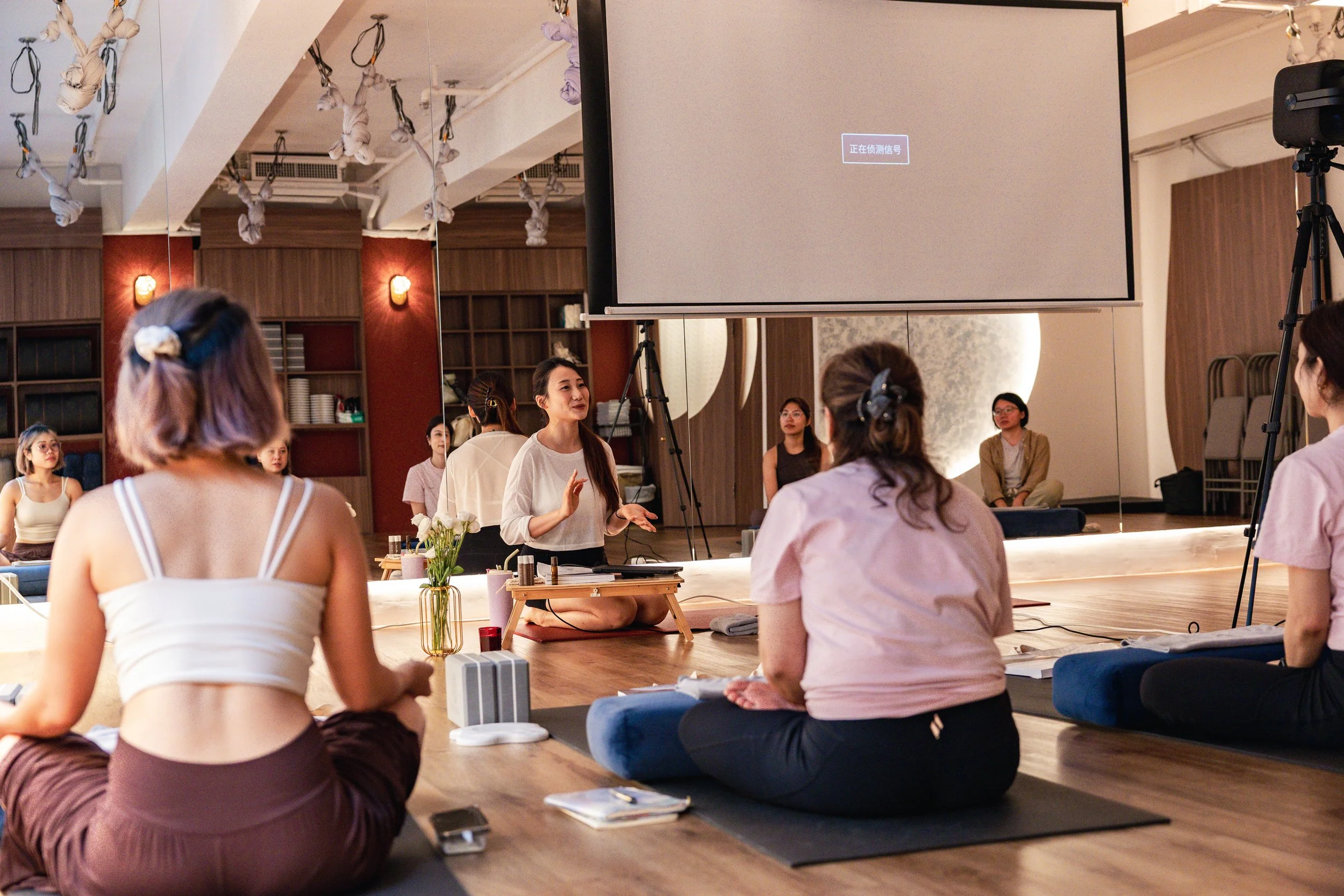 A group of women participating in a yoga or meditation class in a studio, seated on mats in a circle. One woman, possibly the instructor, is talking and gesturing. The room features wooden walls, a large mirror, and a TV screen with Chinese characters that read 'detecting signal.'