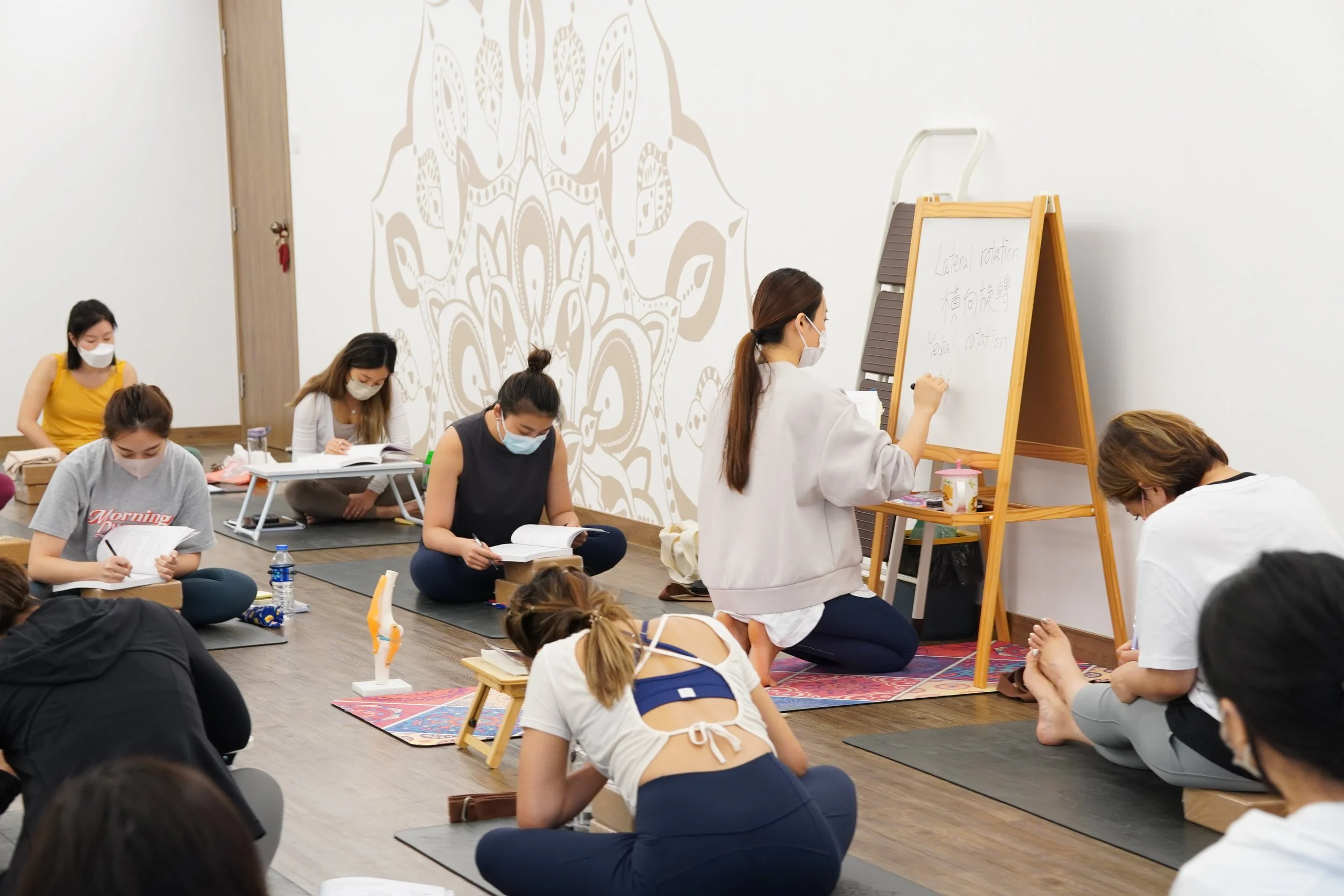 Women participating in a yoga or meditation class, sitting on yoga mats with some taking notes, while an instructor writes on a whiteboard.
