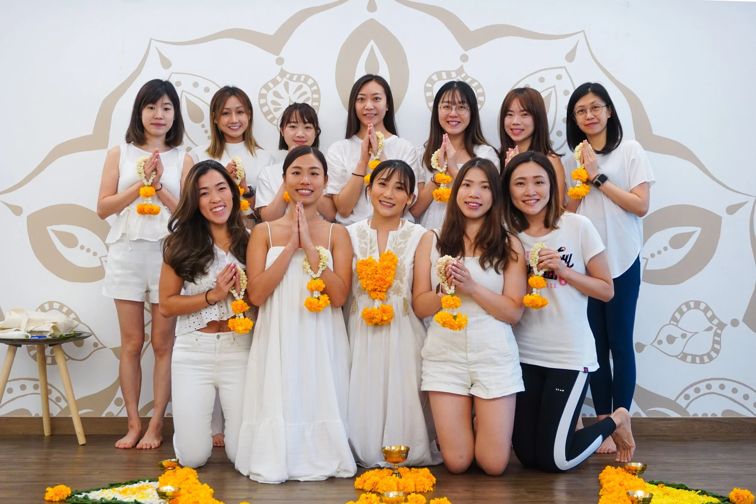 Group of women posing in a cultural celebration, dressed in white, holding flower garlands, with marigolds and offerings on the floor. They are standing and kneeling on a wooden floor in front of a wall with a decorative background.
