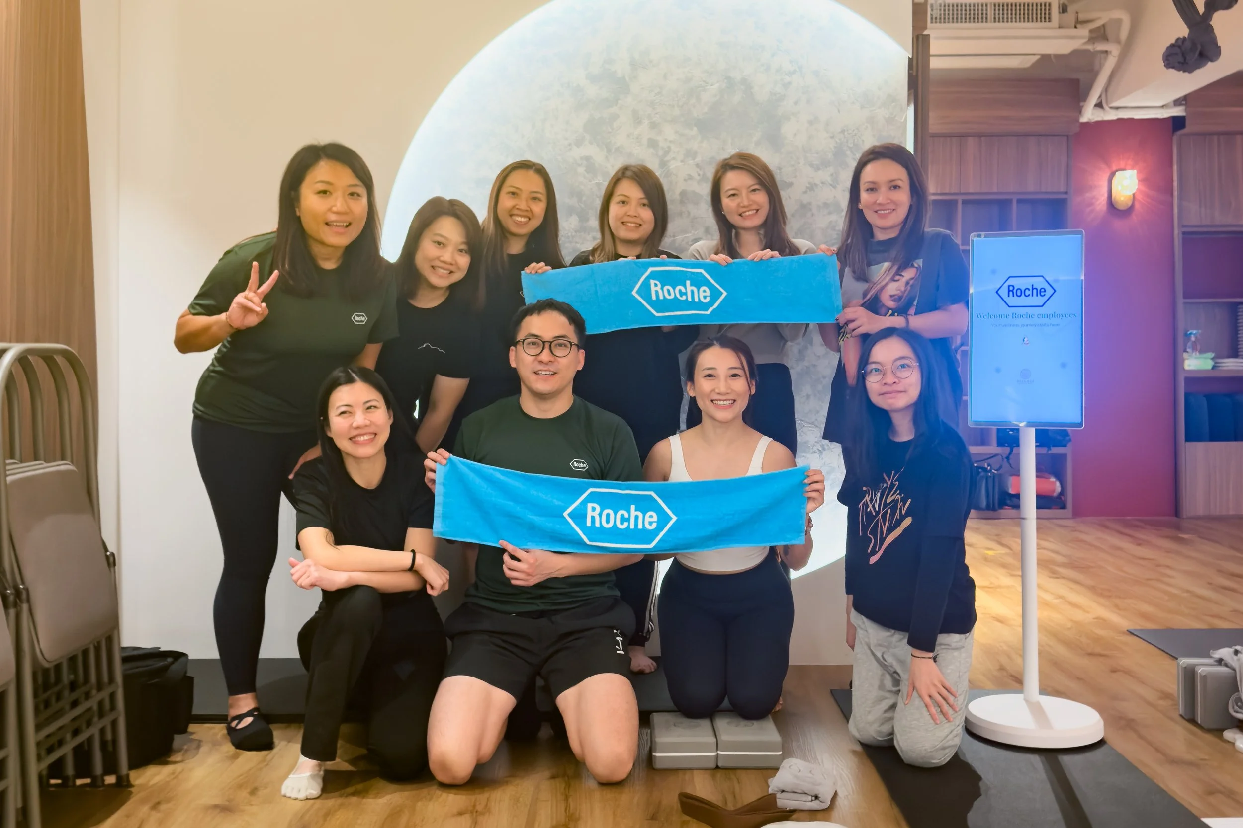 Group of people posing for a photo holding blue Roche banners, inside a room with wooden floors and a decorative wall in the background. They are just done of the Corporate Yoga with Eunice Tsoi, Instructor, at IMAYOGI